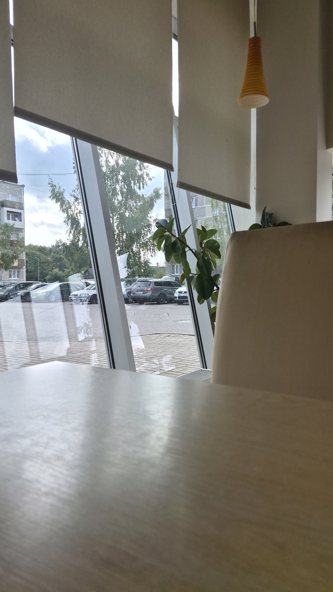 The image appears to be taken from inside a room, possibly a cafe or office, looking out a large window. In the foreground, the surface of a light-colored wooden table is visible, with reflections of light indicating it is polished. Beyond the table, the large window reveals an outdoor scene. The window is divided by angled white frames.
Outside, a parking lot is visible with several cars parked. The cars appear to be dark-colored sedans and SUVs. Beyond the parking lot, there are trees with green foliage and buildings, which seem to be apartment blocks. The sky is cloudy with patches of blue. Roller blinds are partially drawn on the window, obscuring some of the view. A single potted plant with green leaves is positioned to the right of the window, and a portion of a light-colored chair is also visible on the right side of the frame. A distinctive orange lampshade hangs from the ceiling on the right side of the image.
Based on the presence of apartment buildings and a parking lot, the scene could be located in a residential or mixed-use area within the city of Ludza, Latvia. However, without further context or identifying landmarks, it is impossible to pinpoint the exact location.