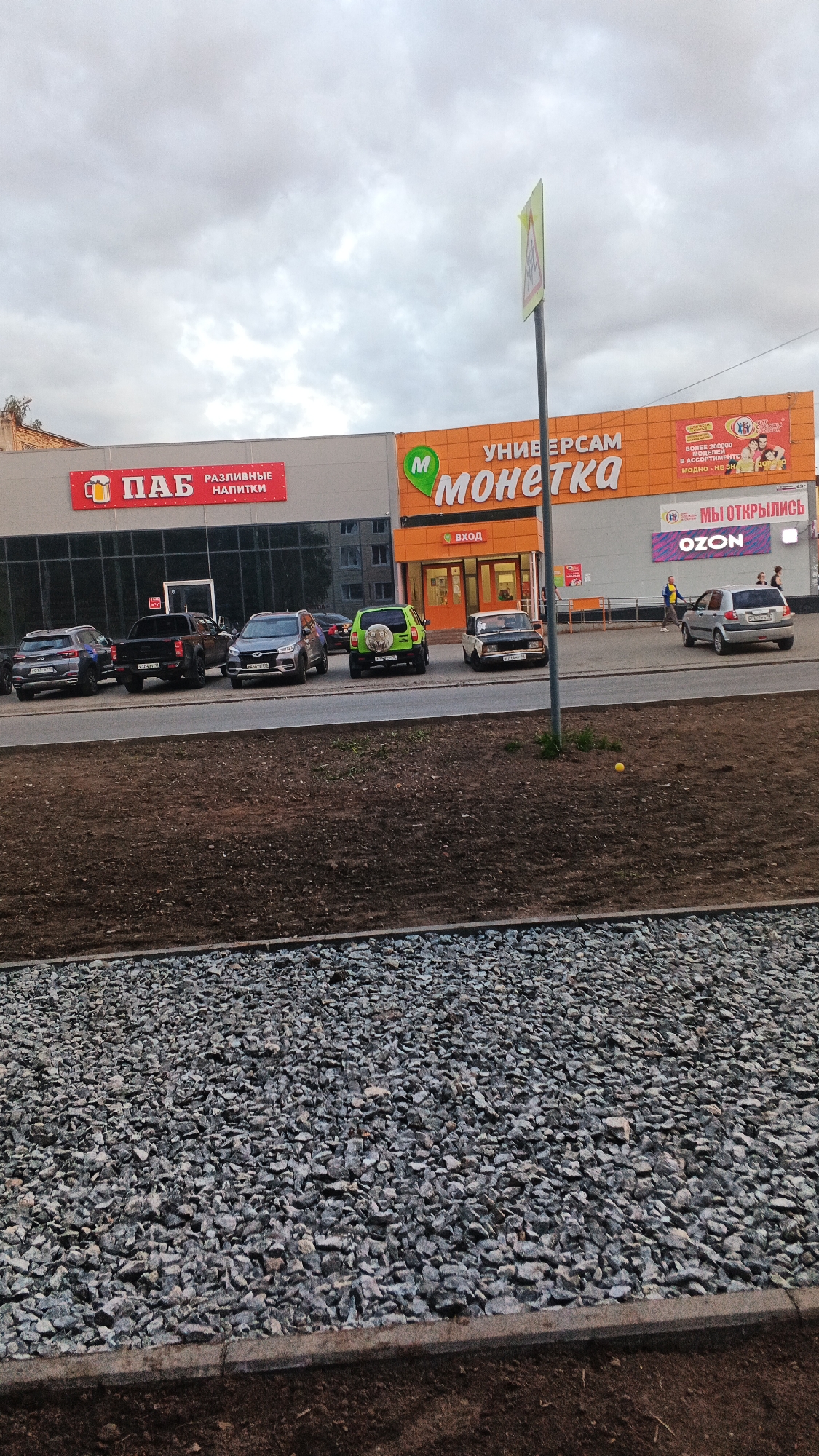 This image shows a street view in Izhevsk, Russia, featuring a row of storefronts along a street. The buildings display prominent signage for "ПАБ" (Pub) and "УНИВЕРСАМ Монетка" (Supermarket Moneta). There is also an "OZON" logo visible, indicating a presence of the online retailer's pick-up point. Several cars are parked in front of the establishments. The foreground shows a landscaped area with gravel and dirt. The sky is overcast, suggesting a cloudy day.
