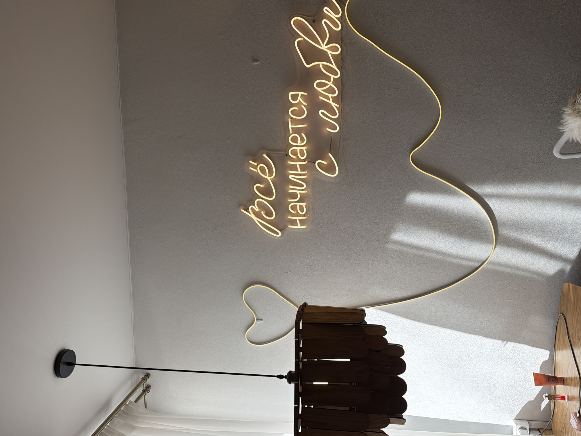 The image shows a room interior, likely a bedroom or living space, with the camera pointed upwards towards the ceiling. A neon sign in Cyrillic script dominates the upper portion of the frame, reading "Всё начинается с любви" ("Everything starts with love"). The neon tubing forms a heart shape around the script. Below the sign, a pendant light fixture with a wood-slat shade hangs suspended. The light is off. Part of a curtain rod and curtains are visible in the bottom left. To the right, a portion of a table or desk and some small items (cosmetics or toiletries) are visible. The location in Votkinsk, Russia, is not directly discernible from the visual content of the image. The location can not be confirmed without additional information.