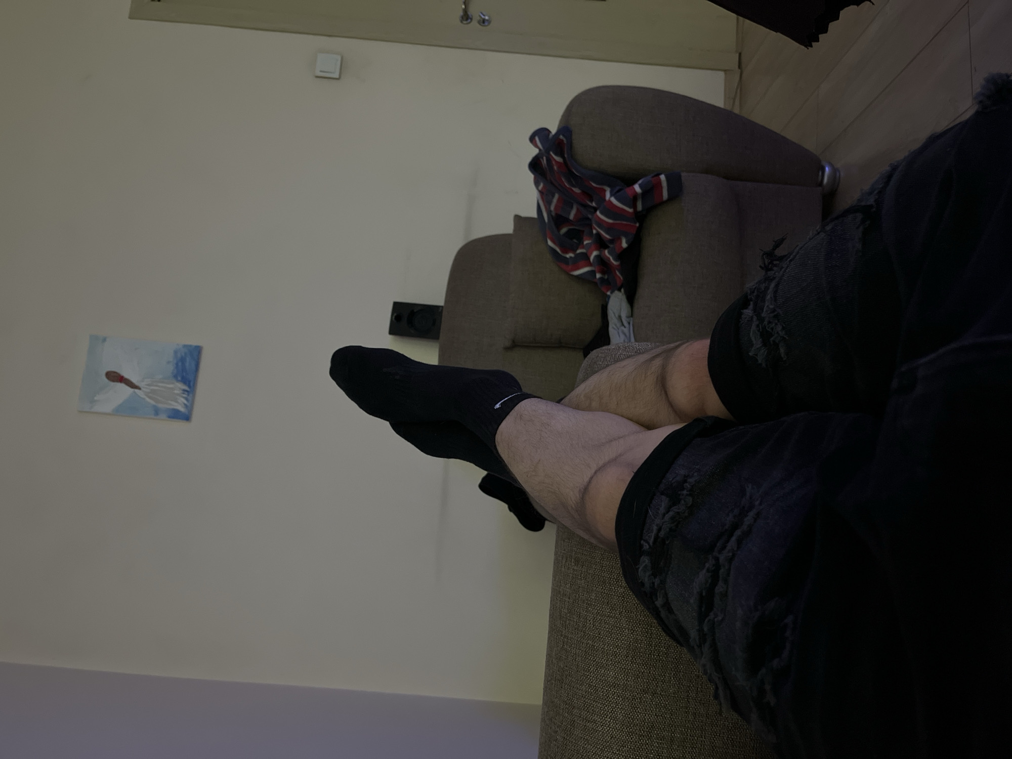 This media file captures an indoor domestic scene, likely a living room or lounge area, in Sofia, Bulgaria, during the evening or night, given the dim, artificial lighting.
The primary subjects are the lower body of a person and a multi-cushioned couch upholstered in a light brown or beige textured fabric. The person's legs are visible, covered in dark, hairy skin, suggesting an adult male. They are wearing black athletic socks, one of which clearly displays a white Nike swoosh logo on the outer ankle. The person is also wearing dark, possibly black, distressed denim shorts or pants with visible rips and frayed edges, indicative of a casual style.
The activity taking place is that of relaxation or leisure, as the person is reclining on the couch, with their feet propped up. The image appears to be taken from the person's own perspective, looking down at their legs, or from a close, slightly elevated vantage point.
Notable details in the setting include an off-white or light beige wall in the background. A small, framed abstract painting hangs on this wall, depicting a red or orange elongated shape over a white and blue background, possibly an interpretation of a figure or object on water. Further up the wall, a white square light switch is visible. A striped shirt, predominantly red, white, and blue, is casually draped over the back and armrest of the couch. On the right side of the image, darker wood paneling can be seen, suggesting a corner or adjacent wall, and a metallic fixture, possibly a door handle, is partially visible. A black rectangular object, likely a speaker or part of an entertainment system, is mounted on the wall near the couch.
The only visible text is the "Nike" brand name on the socks. The overall mood conveyed is one of comfort and informality.