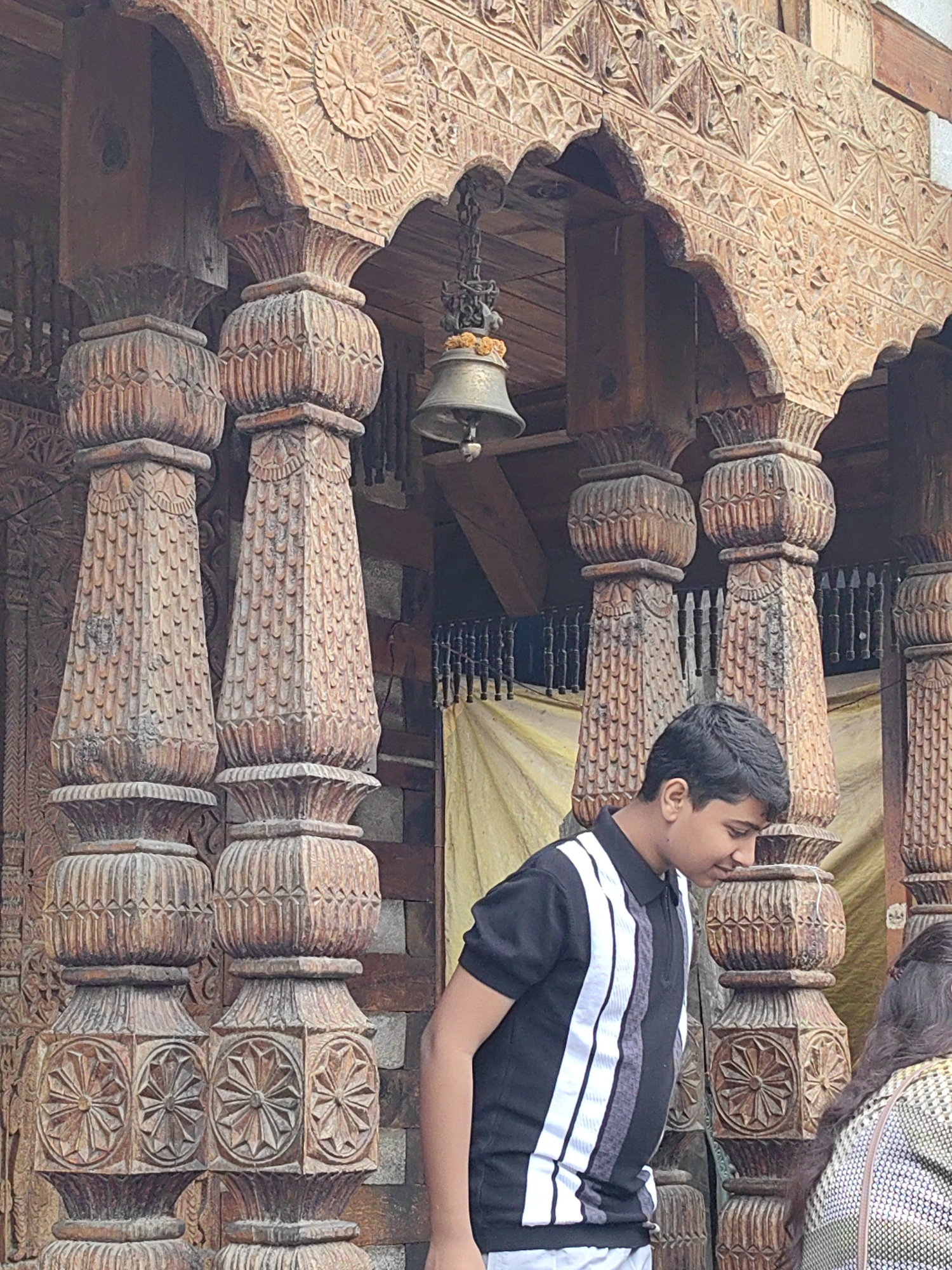 A young boy with dark hair is standing in front of a temple in Vashisht, India. He is wearing a black polo shirt with white and gray stripes. The temple features intricate wooden carvings and columns. A bell hangs from the temple's ceiling. The boy is looking down and smiling. To his right, a woman with long dark hair braided down her back is partially visible, facing away from the camera. The background includes a yellow tarp and more wooden structures.