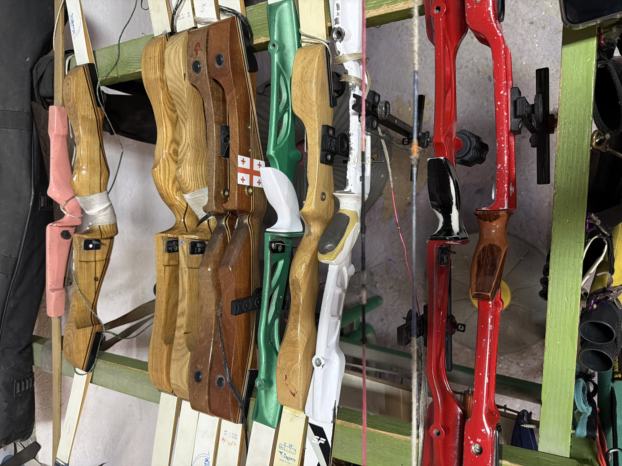 The image displays a diverse collection of recurve bows, primarily risers, meticulously arranged on a simple green wooden rack. The scene appears to be an indoor storage or display area, likely within an archery club or shop in Tbilisi, Georgia, given the array of equipment.
Ten different bows are visible, showcasing a variety of materials, colors, and designs. On the far left, a pink bow riser, possibly made of plastic or composite, is partially seen. Adjacent to it are several traditional wooden risers with natural wood finishes, some featuring white tape or wraps around their grips. One wooden riser prominently displays a small sticker of the Georgian flag – a red cross on a white background. Following these are a distinct bright green riser, another wooden one, and a sleek white recurve riser, likely made from a modern composite material. This white bow has visible "SF" branding and an attached tag showing "28"" and "lbs" measurements, with the brand "Ragim" also faintly discernible. To the right, two vibrant red metal risers are visible, one fitted with a black and white grip.
Many of the bows are equipped with standard archery accessories such as arrow rests and sights. The background is a plain wall, with a black bag partially visible on the far left. No direct activity is depicted; the bows are in a state of organized storage or awaiting use. The lighting suggests a typical interior setting. Visible text includes "Archery" on one tag, "SF," "Ragim," "28"," and "lbs" on other tags, and the distinct Georgian flag.