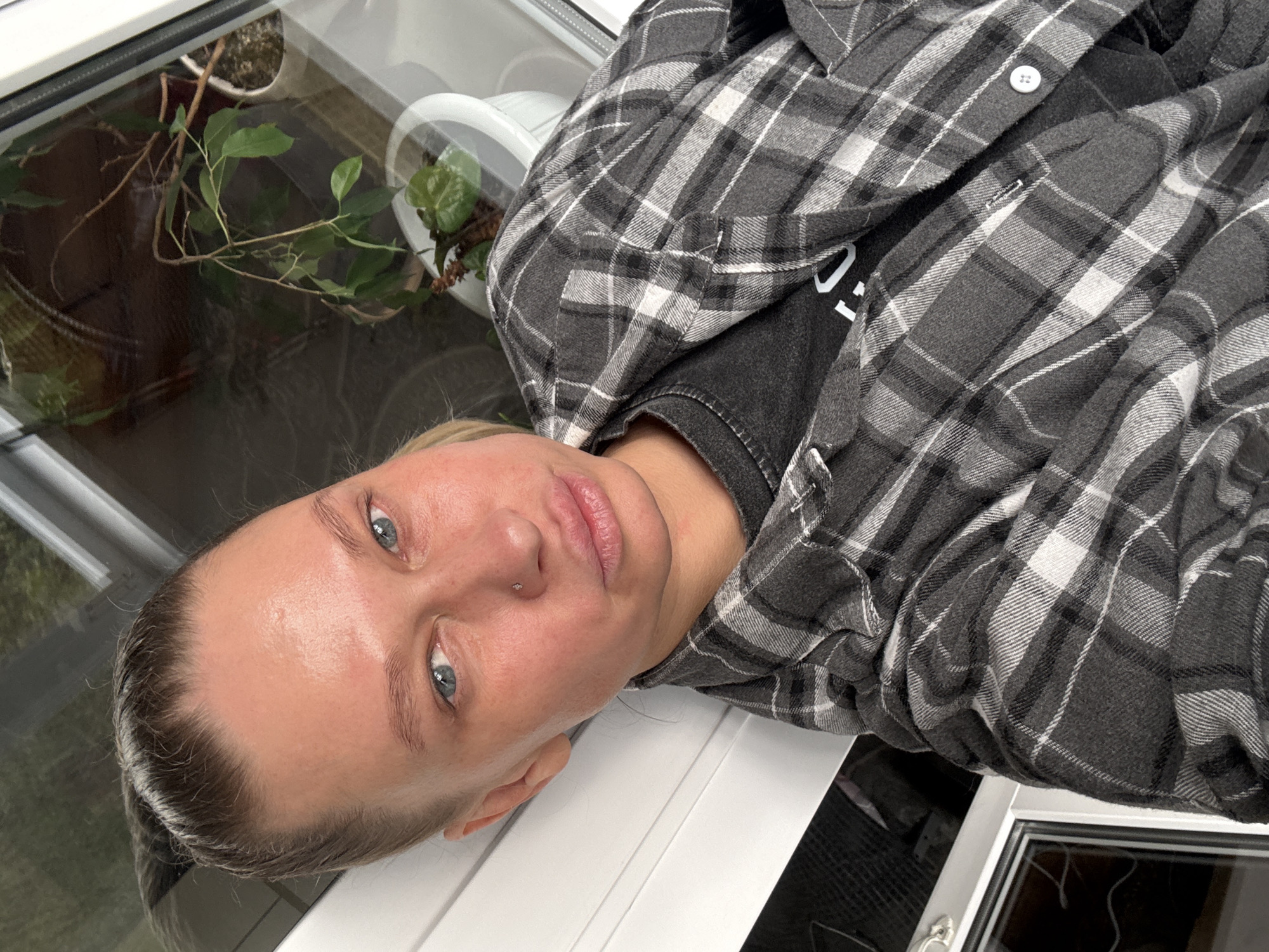 A photograph taken at a low angle depicts a woman with light blue eyes and a fair complexion wearing a gray and white plaid shirt over a dark-colored t-shirt. She has a nose piercing and her hair is pulled back. The background includes plants in white pots, visible through a window, and the white frame of what appears to be a window or door. The precise location in Perm, Russia, cannot be determined from the visual information provided.
