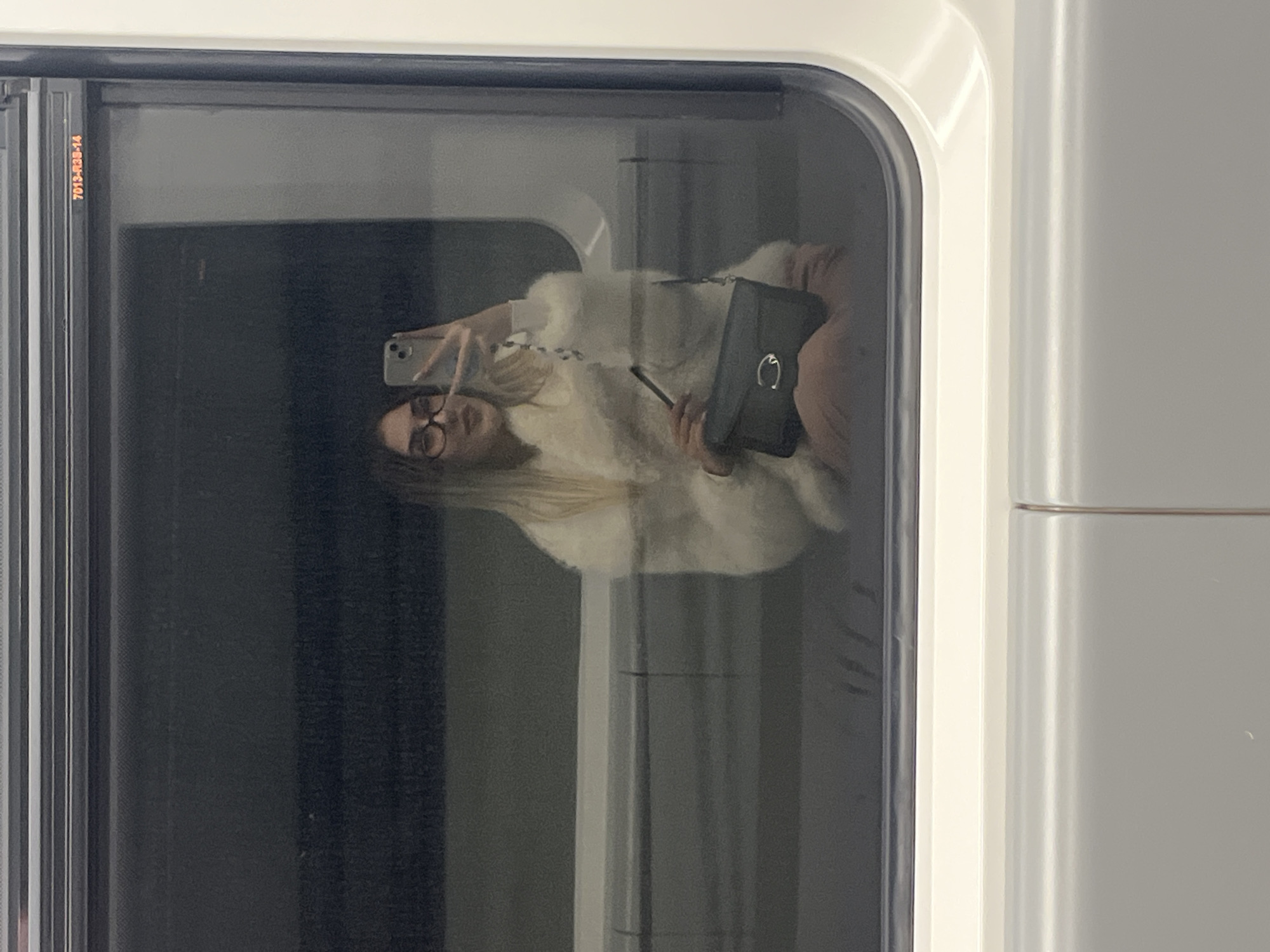 A woman with long blonde hair and glasses is reflected in the window of a train or tram. She is wearing a light-colored, possibly white, furry jacket and is holding a smartphone in one hand and a black handbag in the other. The reflection suggests she is taking a photo of herself. The train or tram window is surrounded by a white frame. The presence of a number "7013-R30-14" suggests the scene takes place in a modern train used in public transportation, potentially in the city of Sofia, Bulgaria.