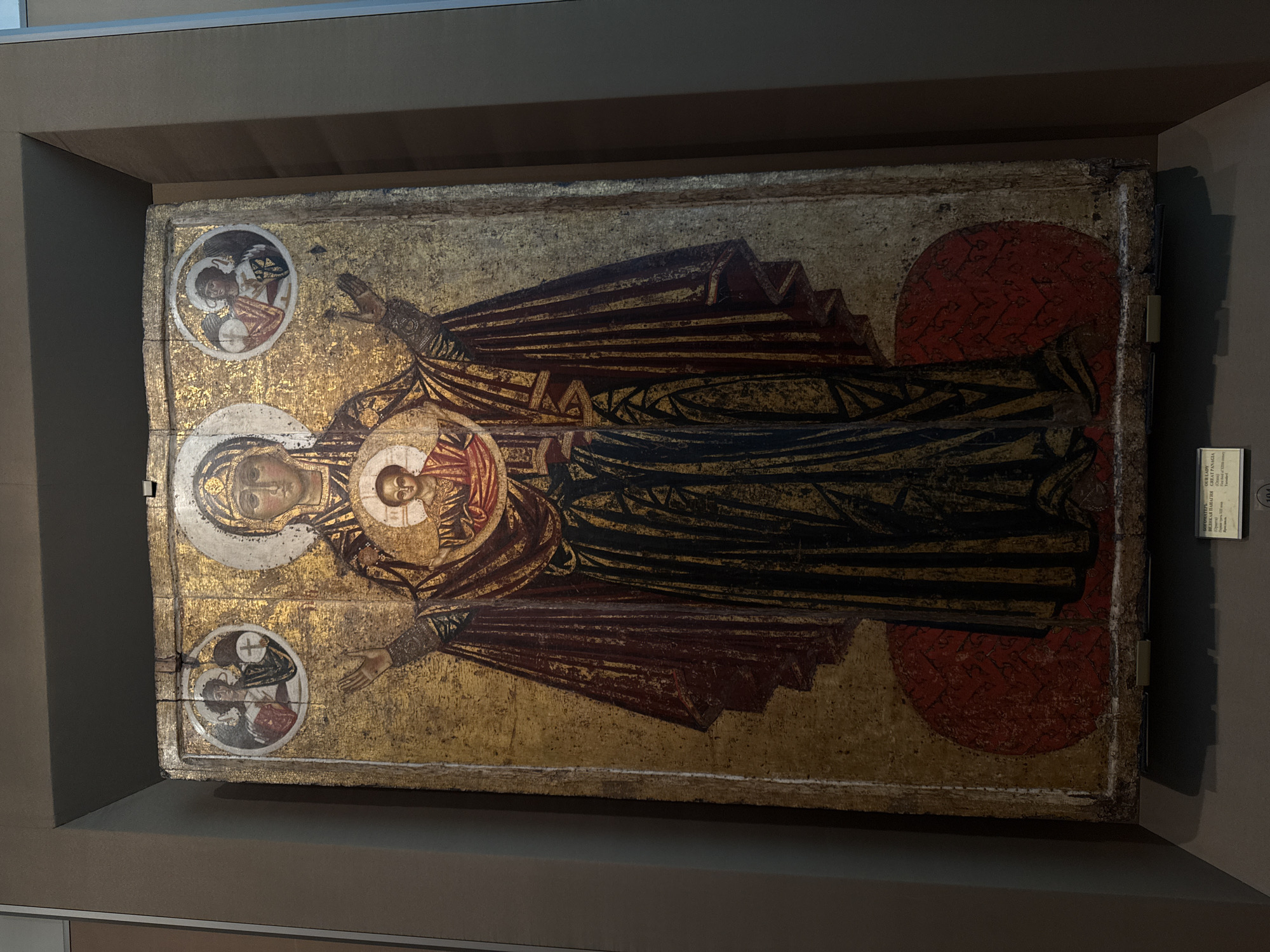 The image shows an Orthodox Christian icon titled "Our Lady of the Great Panagia" (late 12th century). The icon is on display in a museum. The icon depicts Mary holding the Christ Child in a circular medallion in her chest. Her arms are outstretched. Two circular images with religious figures are in the upper left and lower left of the icon. The icon is painted with gold leaf and vibrant colors, particularly red and black. The museum environment includes neutral-toned walls and display cases. The display appears to be in a museum in Moscow, Russia.