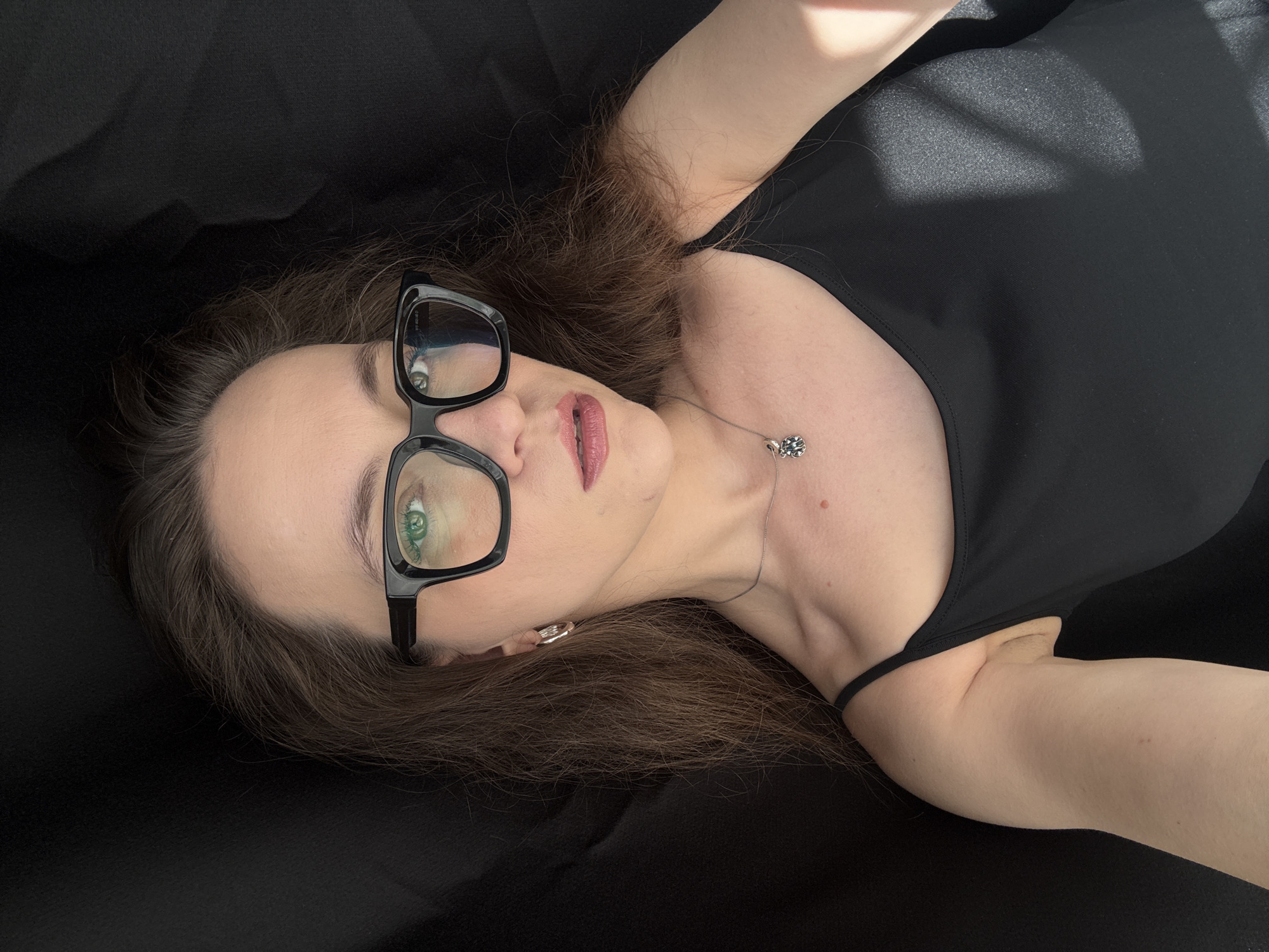 A woman with brown hair and green eyes lies on a black surface, wearing black-rimmed glasses and a black top. She has on a necklace with a silver pendant. The image is illuminated by a strong light source from the top right, casting shadows on her body and the fabric. The woman's face is angled towards the camera, and she appears to be looking directly at it. Her lips are slightly parted, and she is wearing lipstick. The overall impression is a casual, self-portrait. The location is identified as Chaikovsky, Russia. There is no discernible text in the image.