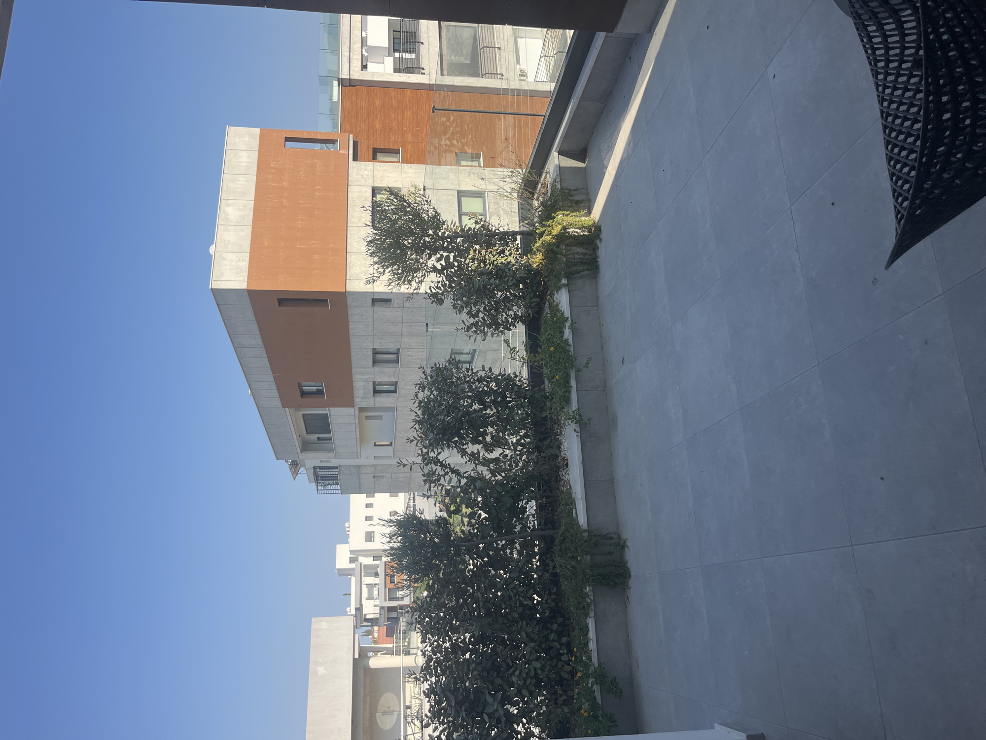 The image shows a view from a tiled patio overlooking a modern apartment building and other residential structures in Agios Athanasios Municipality, Cyprus. The patio has a raised planter box filled with dense green bushes and a small tree. The apartment building features a distinctive color block design with sections of concrete and orange-brown textured material. Balconies with glass railings are visible. The sky is clear and blue. The scene appears to be captured during daylight hours.