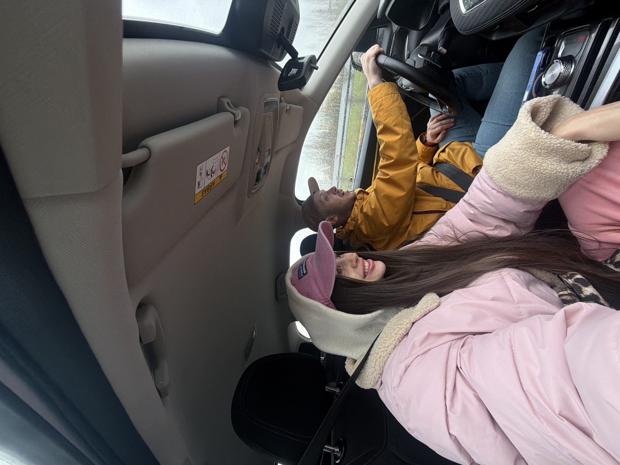 The interior of a car is depicted, with a man driving and a woman in the passenger seat. The man is wearing a yellow jacket and a brown hat, gripping the steering wheel. The woman is wearing a pink jacket, a pink hat, and has long brown hair. The car's dashboard and interior features are visible. The setting does not depict or provide details that would identify a location as Saint Petersburg, Russia. It shows the interior of a vehicle, not an external environment.
