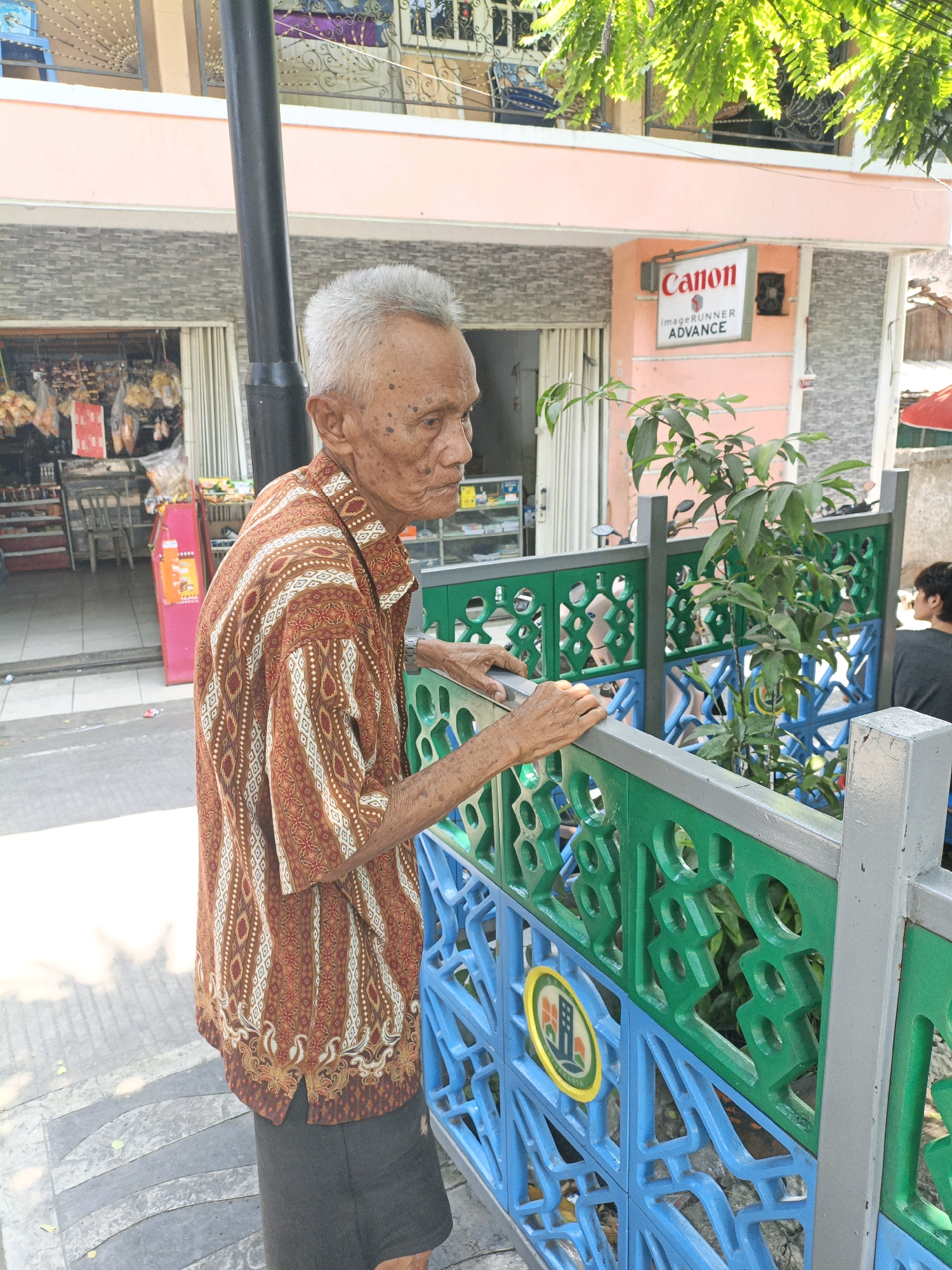 An elderly Indonesian man with graying hair and a patterned short-sleeved shirt is standing on a sidewalk, leaning on a green and blue decorative fence. He is looking to his right, his face partially in shadow. Behind him, a building with a pink facade has a sign that reads "Canon imageRUNNER ADVANCE." The building appears to be a shop or business. A small tree with green leaves grows behind the fence. The sidewalk is made of gray and white patterned tiles. The overall scene suggests a street in a city, possibly in the Grogol Petamburan district of West Jakarta, Indonesia.