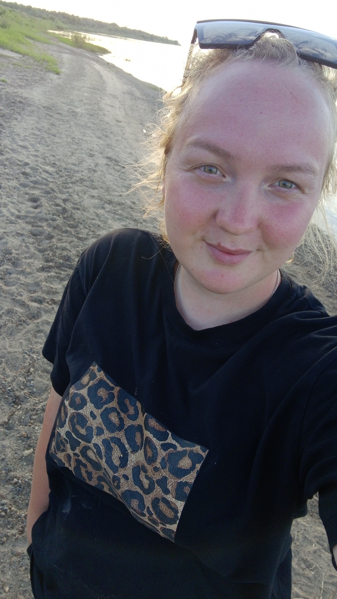 A female subject is captured in a close-up selfie outdoors in Шолья, Russia. She has light-colored, possibly blonde, hair that appears windswept and is pushed back from her face, with strands catching sunlight and creating a halo effect. She has fair skin, flushed cheeks, and green eyes that look directly into the camera. Small lines are visible on her forehead, and a faint smile is present. Dark-colored sunglasses are perched on top of her head. She wears a black short-sleeved t-shirt with a rectangular leopard print patch, featuring brown and black spots, on the upper left chest area. A thin silver-colored chain is visible around her neck.
The background shows a sandy or gravelly ground in the foreground, transitioning to a body of water, likely a river or lake, in the mid-ground. The water reflects bright sunlight, indicating a low sun angle. Beyond the water, a distant landmass with green vegetation and trees is visible under a light sky. The overall environment suggests a natural shoreline or riverbank setting during daylight hours, possibly late afternoon or early morning.