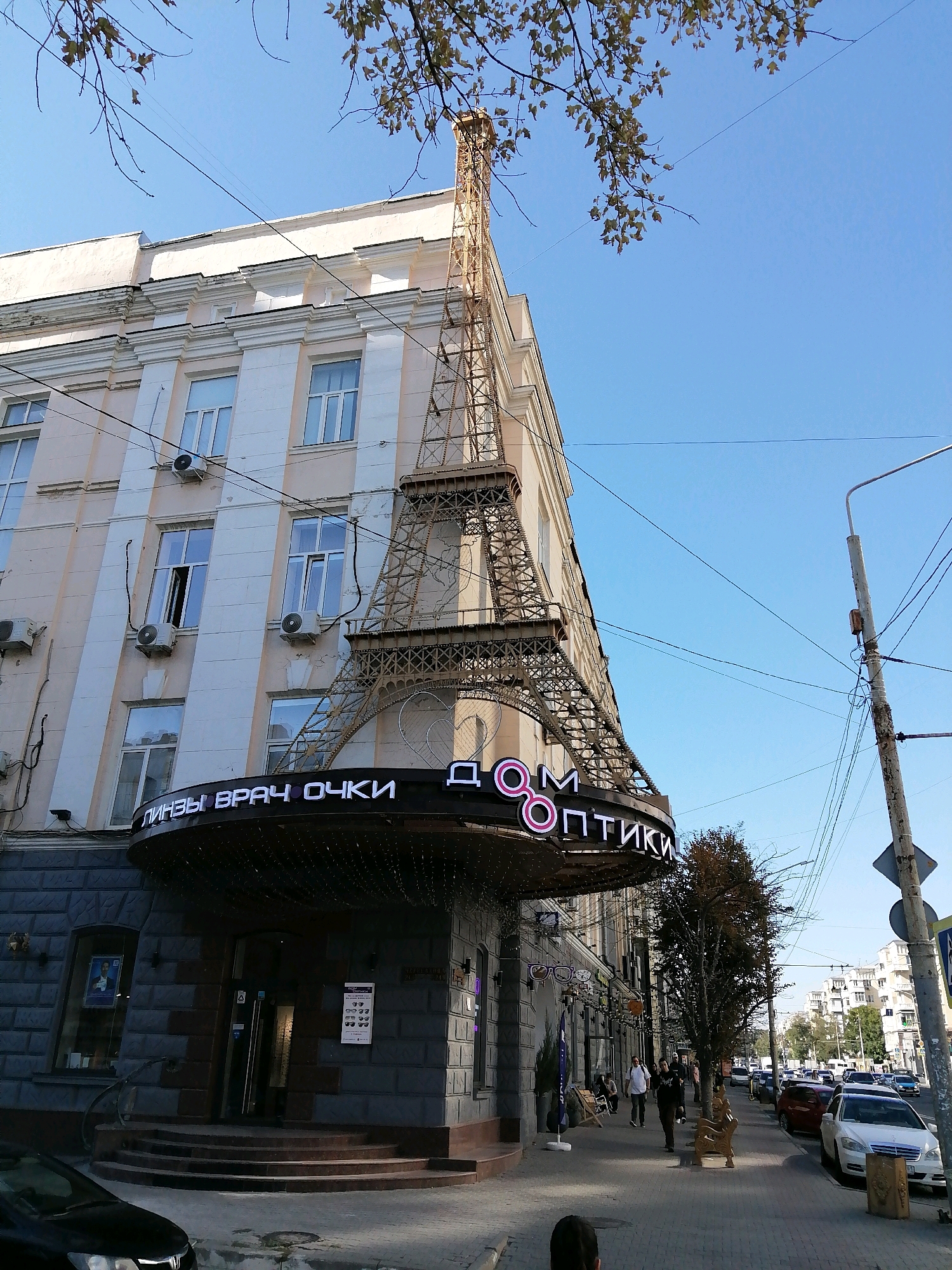 The image depicts a street scene in Rostov-on-Don, Russia, focusing on the facade of a building that features an Eiffel Tower-like structure. The building is several stories high with a pale pink and cream-colored exterior, accented with white decorative molding. Numerous windows, some with air conditioning units, are visible on the upper floors. A prominent sign with illuminated Cyrillic lettering, reading "ЛИНЗЫ ВРАЧ ОЧКИ" (LENSES DOCTOR GLASSES) and "ДОМ ОПТИКИ" (OPTICS HOUSE), is mounted below the Eiffel Tower replica, indicating the presence of an optician's shop. The Eiffel Tower replica is a metallic structure extending from the building's facade.
In the foreground, the entrance to the building is visible, with a dark gray stone facade, a curved staircase leading to double doors, and signage with images of eyeglasses. To the left of the entrance, a black car is partially visible. A sidewalk runs along the building, with a few trees and people walking. A street light and numerous overhead wires are also present. Further down the street, there are cars parked and a line of buildings visible in the background. The sky is clear blue.