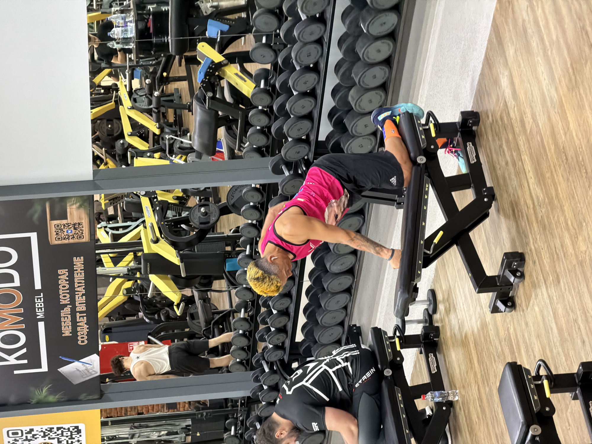 This image depicts the interior of a gym, likely located in Chaikovsky, Russia, given the Cyrillic text on the signage. The gym features rows of dumbbells on racks and various exercise machines, predominantly in yellow and black. In the foreground, a man with vibrantly dyed yellow hair is on his hands and knees on a black weight bench, appearing to perform an exercise. He is wearing a bright pink tank top and black shorts, with colorful athletic socks and shoes. To his left, another man is seated on a black weight bench, wearing a black t-shirt with a white graphic design and black workout pants. He is looking down at his phone or device. In the background, a person in a white tank top and black shorts is standing near some gym equipment. The environment is well-lit, with a polished wooden floor. A sign with the text "KOMODO MEBEL" and a slogan in Russian, "МЕБЕЛЬ, КОТОРАЯ СОЗДАЕТ ВПЕЧАТЛЕНИЕ" (Furniture that creates an impression), is visible in the upper left portion of the frame, suggesting a potential sponsorship or a related business.