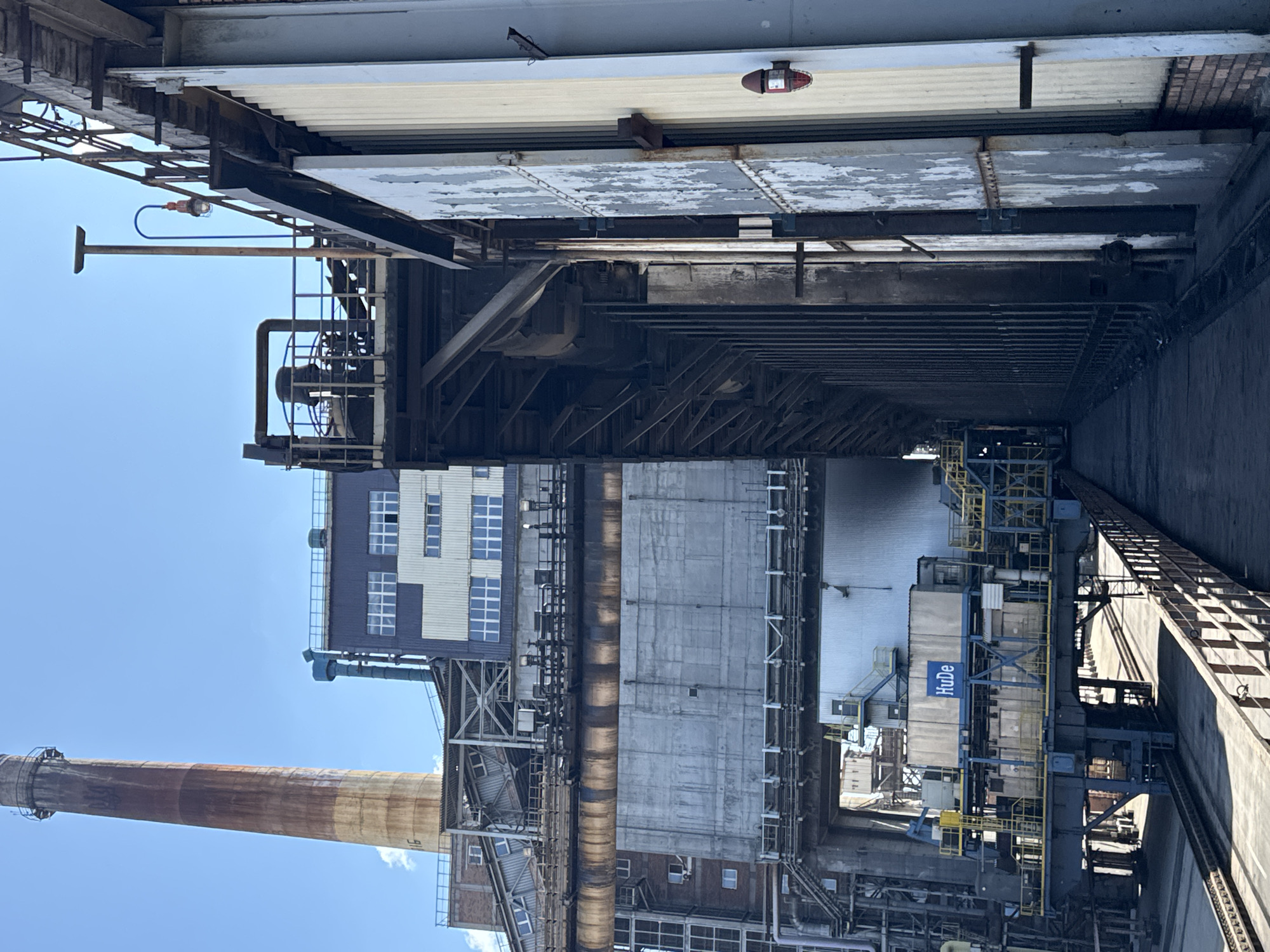 The image depicts a view of an industrial facility with a prominent smokestack in the background. The smokestack is tall and cylindrical, exhibiting signs of rust and weathering. Adjacent to it are various industrial buildings and structures, characterized by metal frameworks, concrete walls, and numerous pipes and conduits. One building has a facade with multiple windows arranged in a grid pattern. In the foreground, there is a metal walkway or platform that runs alongside a large, imposing industrial structure. This structure appears to be a furnace or a similar industrial processing unit, with vertical metal panels on its exterior. On one of these panels, a red warning light is visible. The ground is paved or covered with industrial debris. The sky is clear and blue. The image is taken outdoors on a sunny day, likely within the city of Kryvyi Rih, Ukraine, given the context.