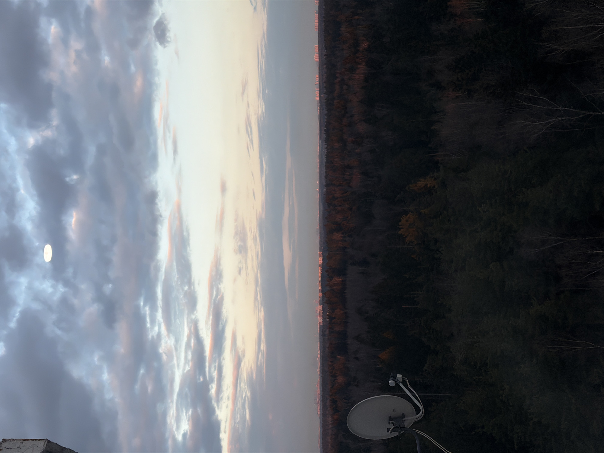 This image presents a panoramic landscape view at twilight, likely evening, from an elevated vantage point.
The sky, occupying the upper half of the frame, is filled with a dynamic array of clouds. These clouds display a gradient of colors, transitioning from soft grey-blue in the upper left to warmer pink, orange, and light grey tones towards the horizon, suggesting the last light of dusk. A prominent, bright, circular white orb, strongly resembling a full moon, is visible in the upper left quadrant, partially obscured by the clouds.
A distinct horizon line separates the expansive sky from the terrestrial landscape. Along this horizon, a faint strip of distant buildings or structures can be seen, their forms dimly lit, indicating a distant town or city in Brekhovo, Russia.
The lower half of the image is dominated by a vast, dense forest that appears largely silhouetted and dark against the lighter sky. The trees are predominantly evergreen, with some bare branches visible, adding to the dark, rich texture of the foliage.
In the lower right corner, a white or light grey satellite dish with its LNB and supporting arms is clearly visible, a modern technological element contrasting with the natural landscape. A small portion of a light-colored concrete structure, possibly a rooftop or balcony edge, is visible in the lower left corner, indicating the photographer's elevated position.
There is no discernible activity or event taking place, and no people are visible. The overall mood conveyed is one of peacefulness and vastness, characteristic of a twilight scene over a natural landscape bordering a distant settlement. No visible text is present in the image.