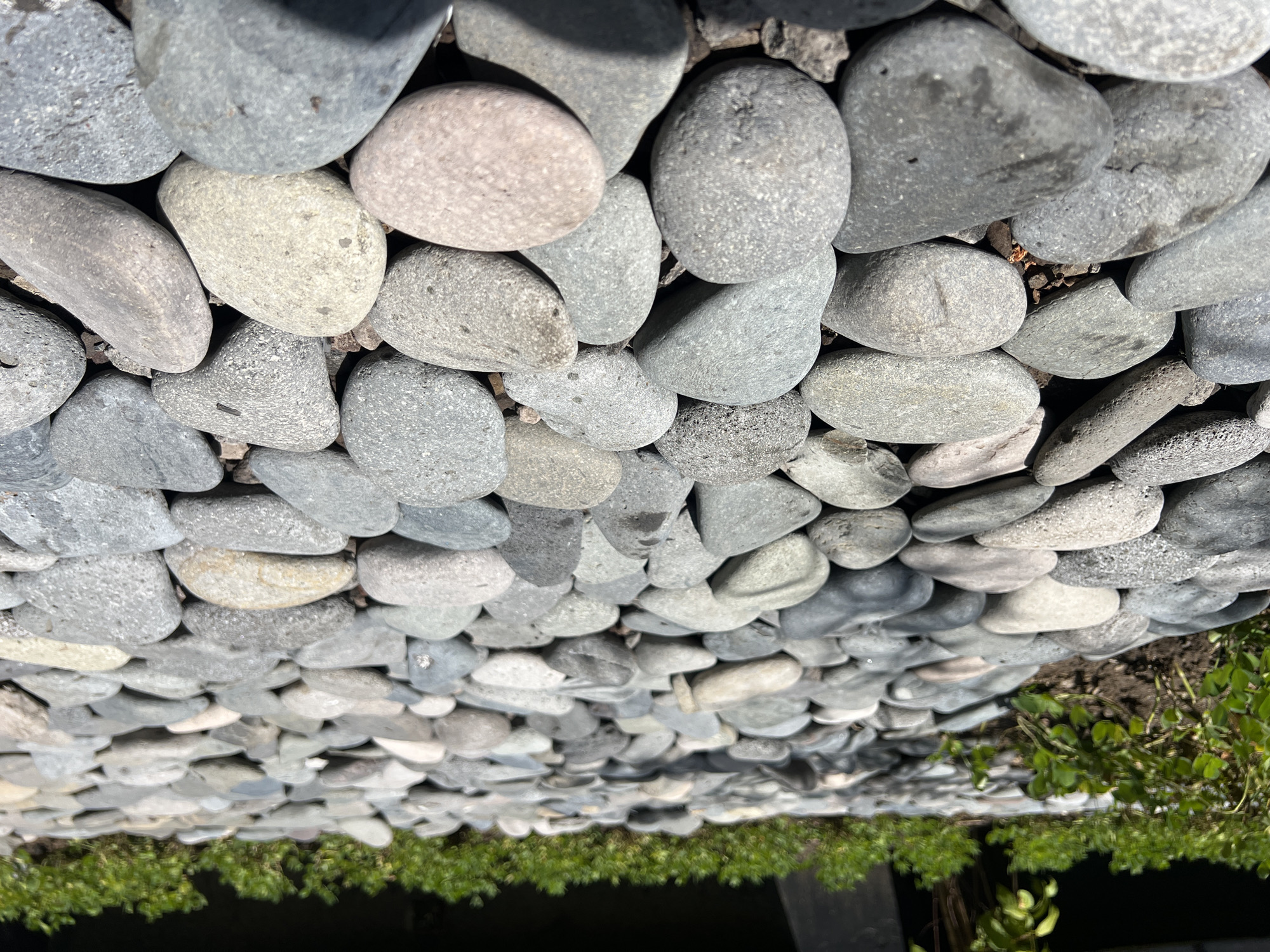 The image shows a ground cover of smooth, grey river rocks, interspersed with patches of exposed soil and green vegetation. The rocks vary in size and color, ranging from light grey to dark grey. The vegetation appears to be small shrubs or ground cover plants. The lighting suggests an outdoor environment. The exact location in North Kuta, Indonesia, cannot be determined from the image alone, but the scene appears to be a landscaped area. No people, specific subjects, actions, or interactions are visible. The objects are rocks, soil, and plants.