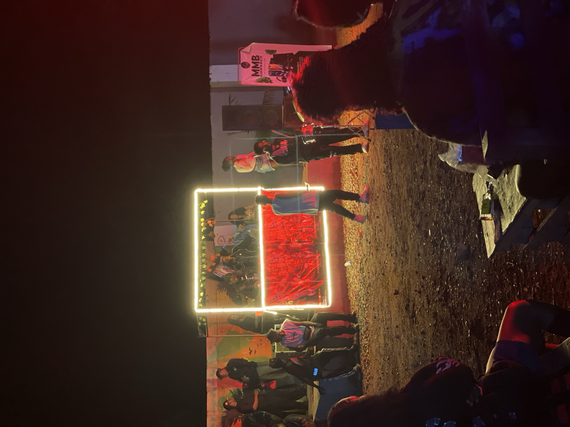 The image depicts an outdoor nighttime event with a stage setup. The stage is illuminated by a prominent neon light frame with a red backdrop. Several people are present on and around the stage, some performing or engaged in musical activities, including what appears to be a drum set and speakers. A banner for "MMB CONNECT" is visible to the right of the stage. A crowd is gathered in front of the stage, some seated and observing the performance. The ground appears to be dirt or gravel. The overall atmosphere suggests a lively event in Durumi, Nigeria.