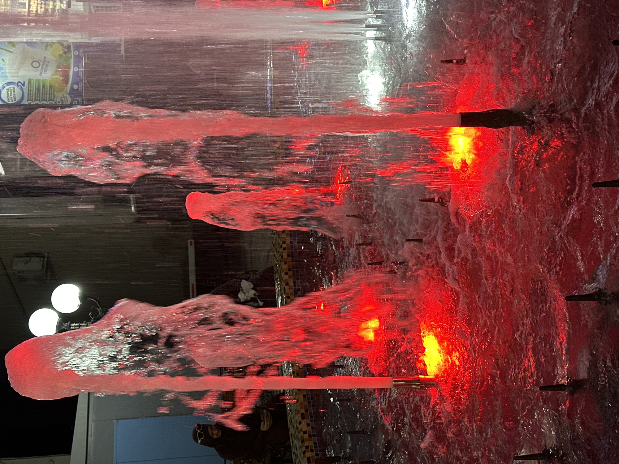 This is a night photograph of a fountain with red lighting located in Tashkent, Uzbekistan. The fountain has multiple jets of water spraying upwards, illuminated by red lights at their base. The water appears frothy and turbulent. In the background, there are lights, buildings, and what appears to be a billboard or advertisement. A person is visible in the left background, seated and wearing sunglasses and a hooded jacket. The overall scene is dark, with the bright red lighting of the fountain being the dominant visual element.