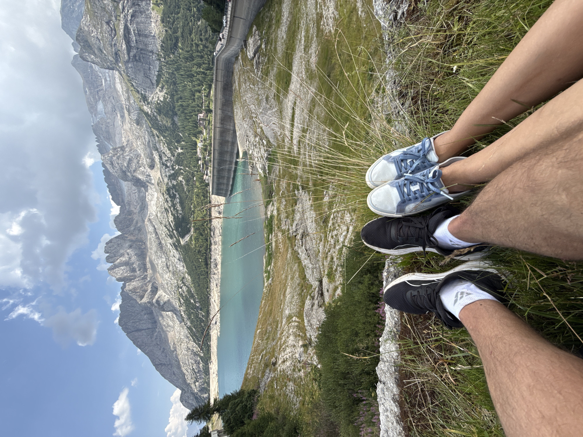 The image shows a scenic view of a mountain landscape with a large reservoir dam. In the foreground, two pairs of legs are visible, suggesting two people are sitting and relaxing. One person is wearing black athletic shoes and white socks, while the other is wearing light blue sneakers. The background features a vast body of turquoise water, a concrete dam structure, a winding road with vehicles, and rugged, grey mountains under a partly cloudy sky. The terrain leading up to the dam is a mix of grassy slopes and rocky outcrops. This location is suggestive of the Fedaia Dam, a prominent feature in the Fedaia Pass, which connects the provinces of Trentino and Veneto in Italy, near Canazei.