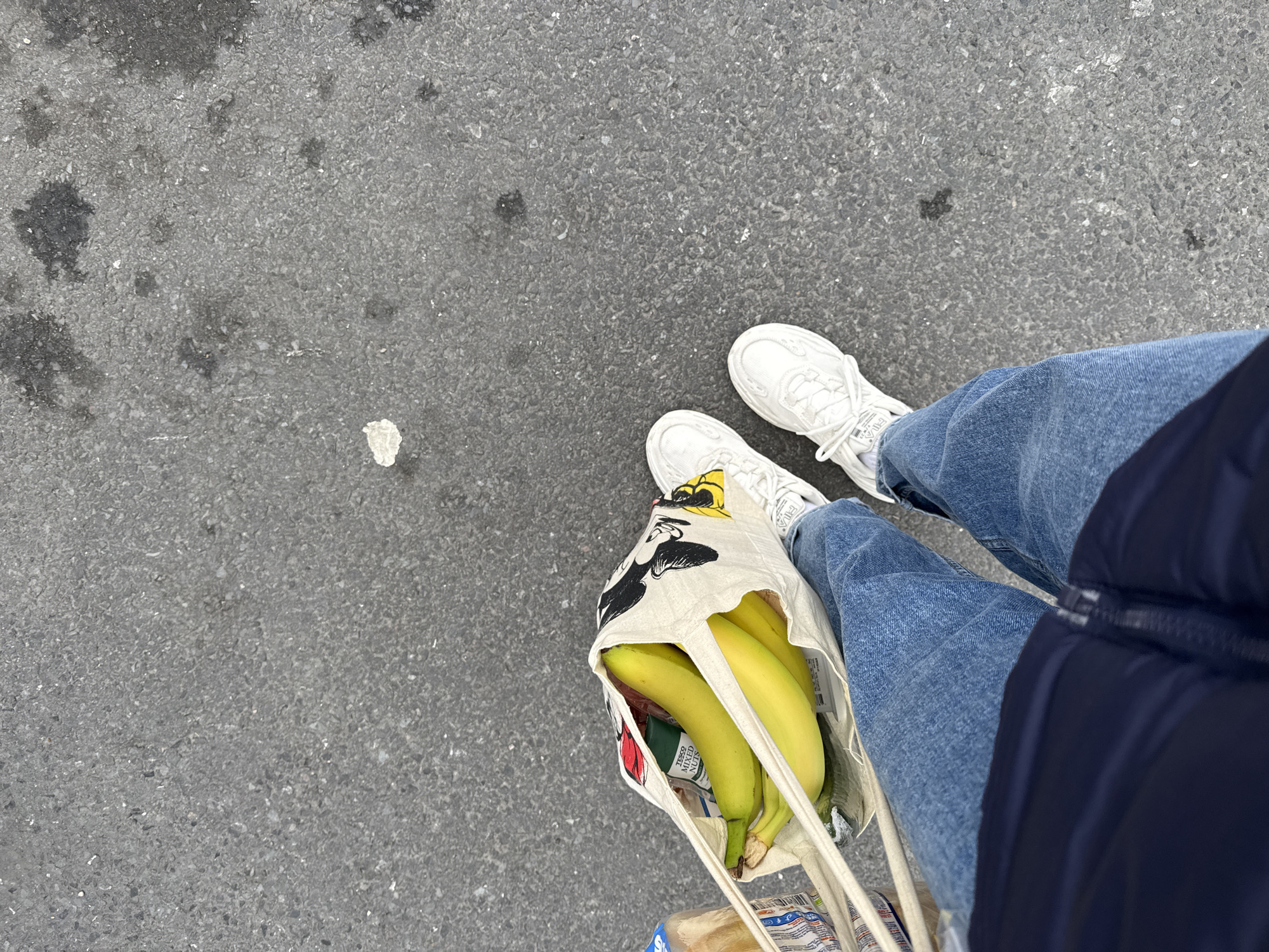 A person wearing blue jeans and white sneakers stands on a grey asphalt surface. They are holding a reusable shopping bag filled with bananas, mixed nuts, and bread. The bag is decorated with a Mickey Mouse pattern. The location appears to be an urban environment, possibly a street in Great Yarmouth, United Kingdom, based on the Tesco brand of the mixed nuts. A portion of a navy jacket is visible on the right.
