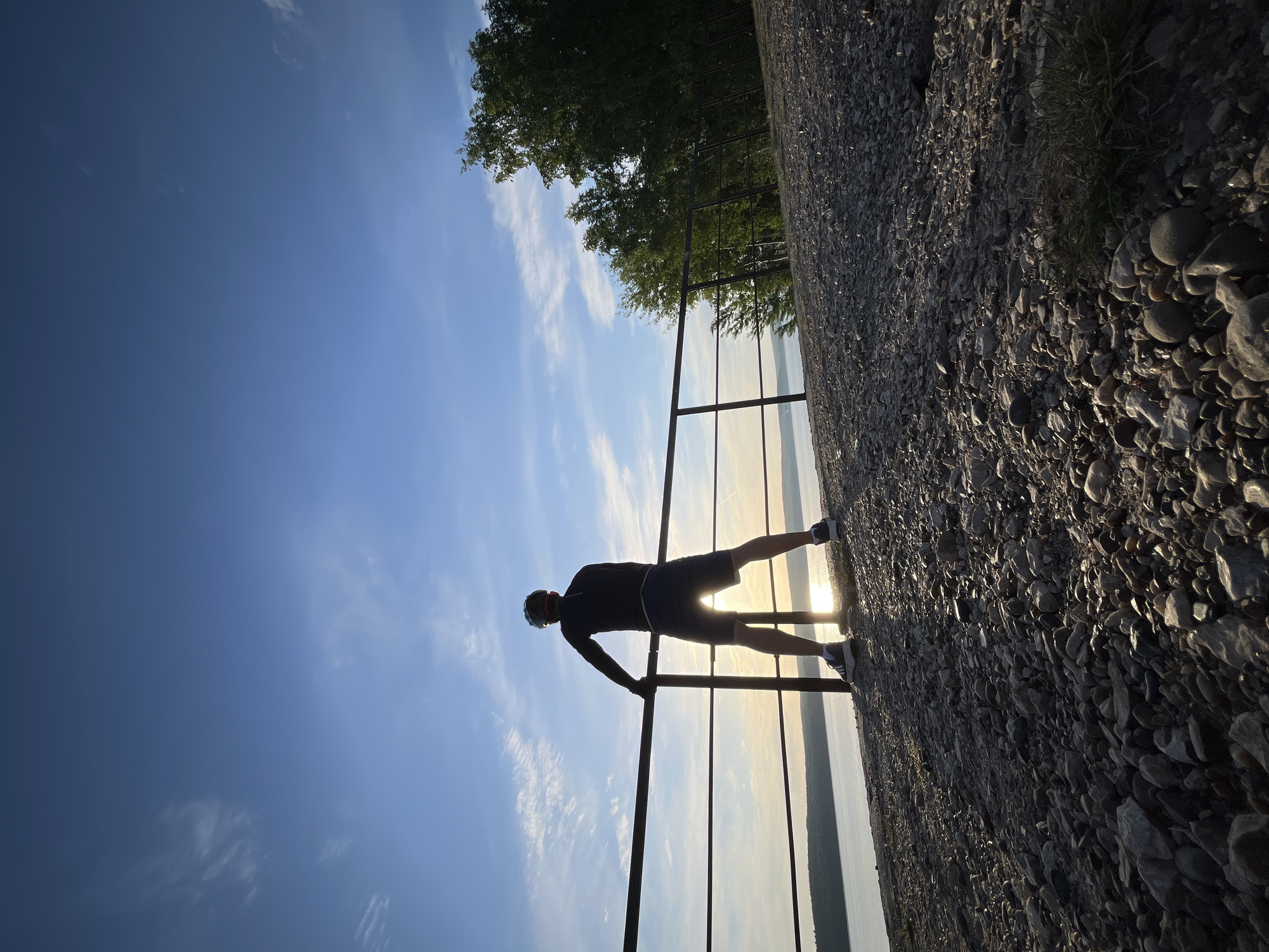 A man's silhouette is captured against a backdrop of a sky transitioning from clear blue to a bright, light-colored horizon, potentially indicating sunrise or sunset. The man is positioned near a metal railing, appearing to either lean on or perform an exercise against it. The ground adjacent to the railing is covered in small rocks or gravel, suggesting a pathway or embankment. Trees are visible in the background. The scene is Chaikovsky, Russia.