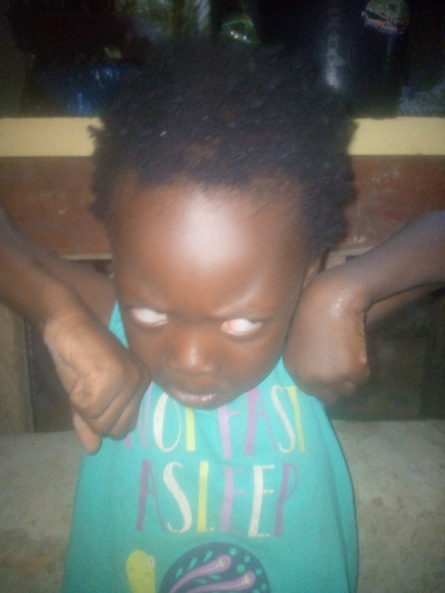 A child is captured in a close-up, low-angle shot. The child has dark skin and short, dark hair. The child is wearing a turquoise-colored top with white and lavender lettering that reads "FAST SLEEP". The child is looking down with their eyes wide open, and arms bent at the elbows. The child's fists are positioned near their face. The background is blurred and suggests an indoor setting, but without clearer details, it's impossible to specify the location in Ikorodu, Nigeria.