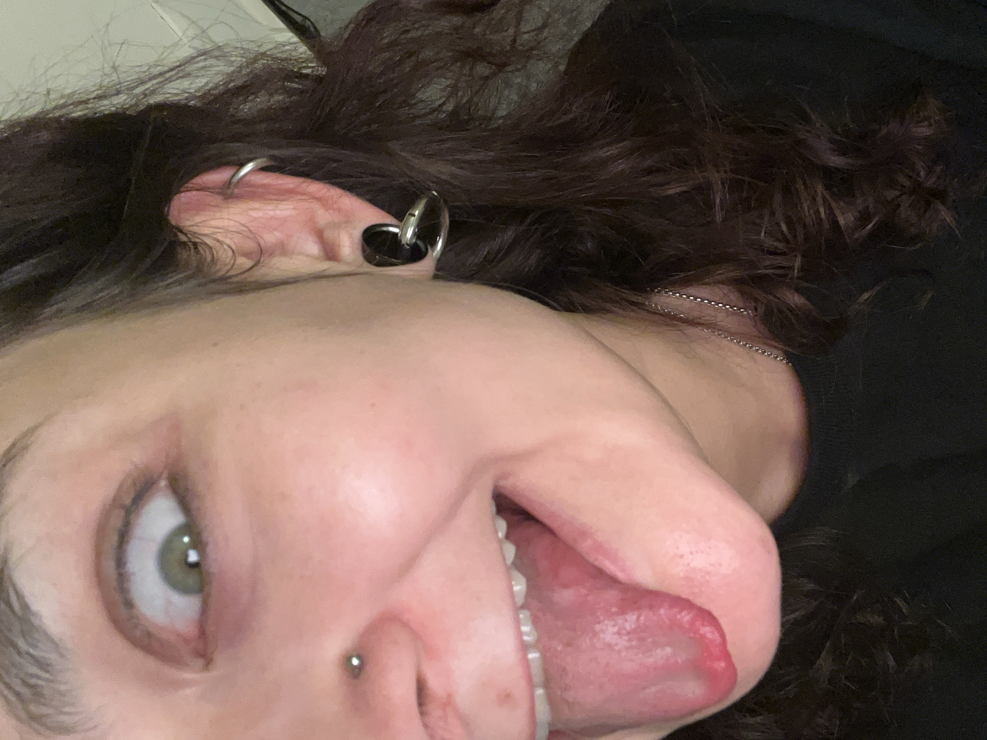 This close-up image features an individual captured in an unconventional, almost upside-down portrait, likely a selfie. The subject's face is centered, displaying one prominent light green eye that directly engages the viewer. Their dark brown, curly hair cascades around their head, blending into an indistinct, dark background.
Notable facial features include a small, silver stud piercing on the side of the nose. The person's mouth is open, revealing white teeth and their tongue playfully sticking out. The expression conveys a sense of lightheartedness or mischief.
The subject's ear is adorned with multiple piercings: a large black and silver circular piece is visible in the earlobe, and a smaller silver hoop piercing is situated higher on the ear. A thin silver chain necklace is also visible around the neck, peeking through the hair. The image is a candid and expressive self-portrait, rich in personal details. No text is visible. The scene appears to be indoors, possibly in Picassent, Spain.