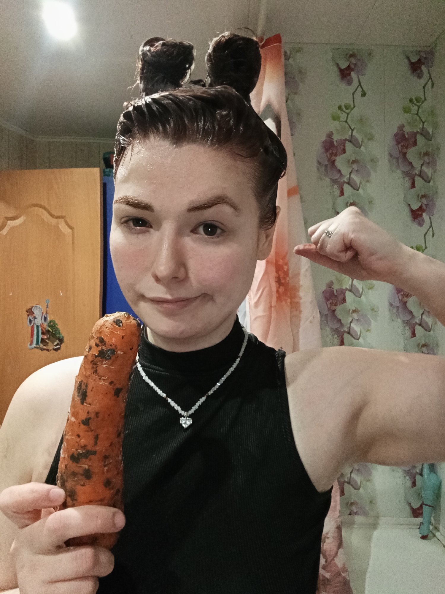 A woman with dark, wet hair styled in two buns on top of her head, wearing a black sleeveless top and a necklace with a heart-shaped pendant, is posing indoors. She is holding a large, dirty carrot in her left hand and flexing her right bicep. The background consists of a light-colored wall with a floral pattern, a light-colored wooden door, and a section of a blue cabinet or shelf. The image appears to be taken in a bathroom or similar domestic setting. The location is possibly in the city of Chaikovsky, Russia, although there is no definitive evidence of this within the image itself.