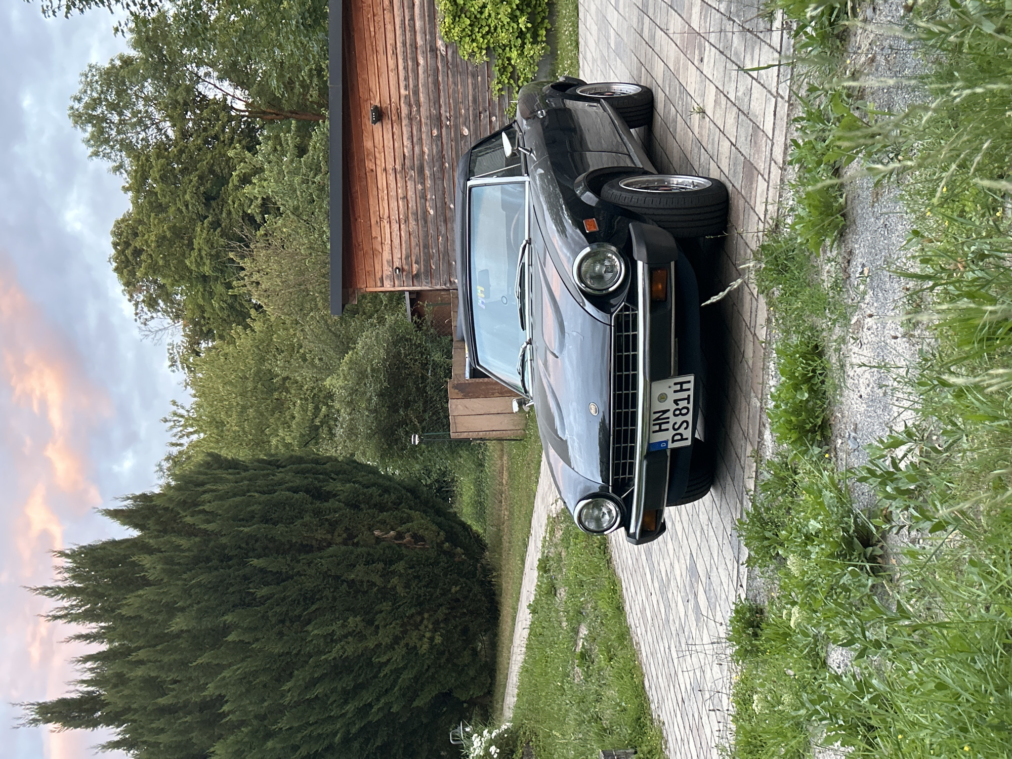 A dark colored convertible Fiat Spider is parked on a paved driveway in front of a rustic wooden building. The car is facing the viewer, angled slightly to the left. It has a black soft top that is up. The car's license plate is visible and reads "HN PS 81H". The driveway is made of light-colored rectangular pavers. To the left of the driveway, there is a path leading away from the car, bordered by grass and some white flowers. Behind the car and building, there is lush green foliage with tall trees and smaller bushes. The sky above is cloudy with hints of pink and orange, suggesting it might be sunset or sunrise. The overall scene appears to be a residential or semi-rural setting.