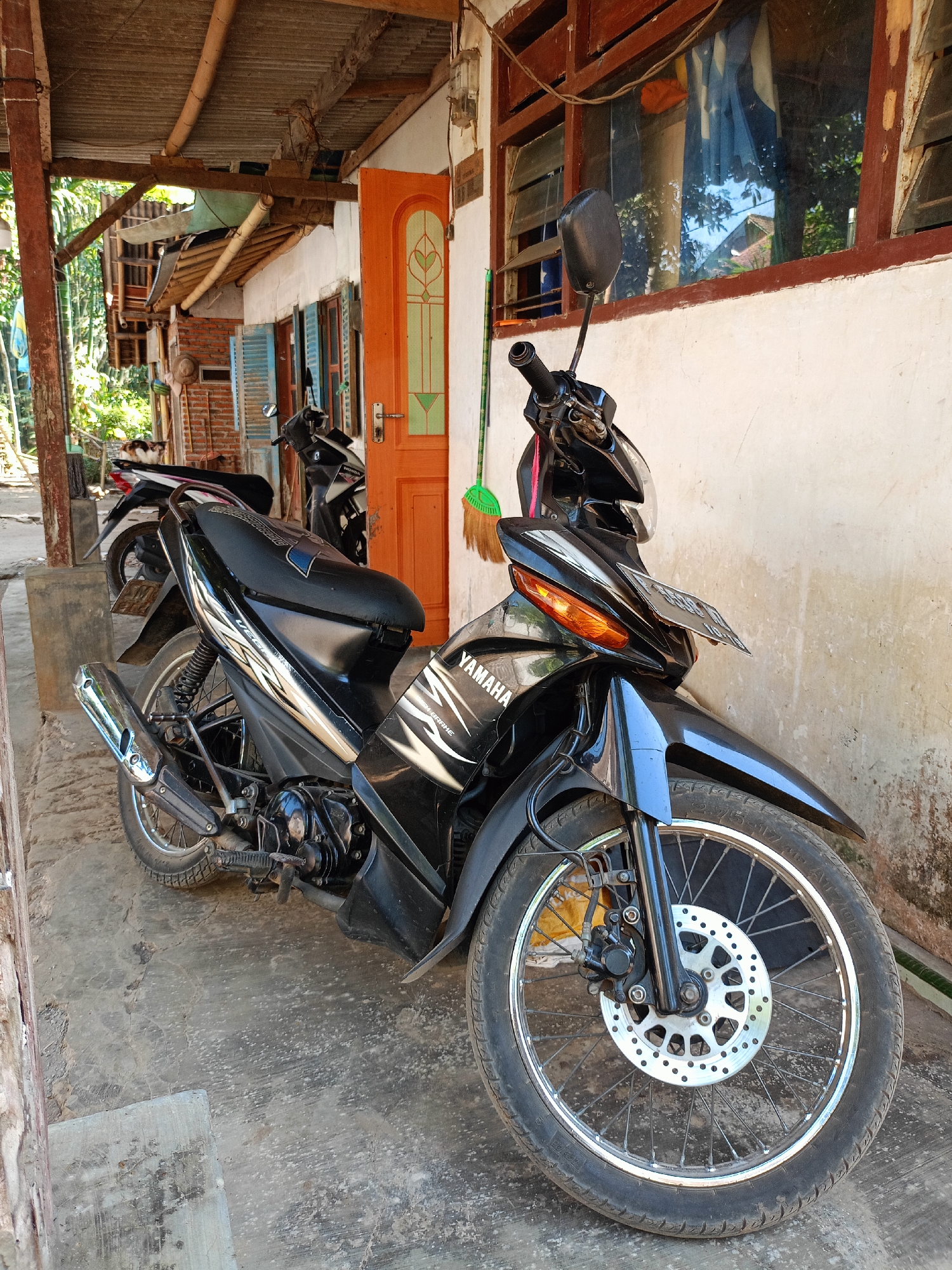 A black Yamaha motorcycle is parked on a concrete ground in front of a building with a wooden roof and orange door. A broom with green bristles is leaning against the wall next to the motorcycle. The building appears to be in a residential area. The image is taken from a medium shot, with the motorcycle as the main subject. The overall lighting suggests it is daytime. There are no people or animals visible in the image. The environment seems to be outdoors, possibly on a street or in a yard. The building has wooden window frames and a light-colored wall. The roof is made of corrugated metal and supported by wooden beams. The scene is set in Jelbuk, Indonesia.