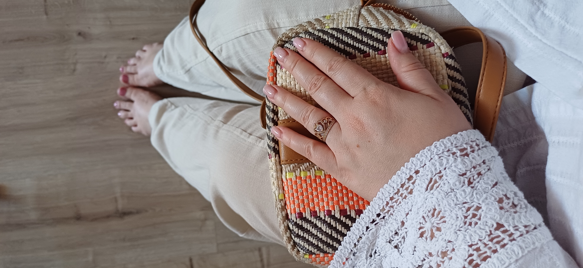 In Sofiivska Borshchahivka, Ukraine, the image captures a close-up of a woman's hand resting on a woven straw bag. The bag features a colorful striped pattern of brown, white, orange, and yellow. The woman is wearing a rose gold ring on her ring finger, which is shaped like a crown and adorned with small stones. Her fingernails are painted a light pinkish nude. She is also wearing a white, crocheted garment with intricate patterns on her forearm. In the background, a person's legs are visible, wearing light-colored pants, and bare feet are positioned on a light brown wooden floor. The scene suggests a casual setting, possibly indoors or on a patio, given the wooden floor.