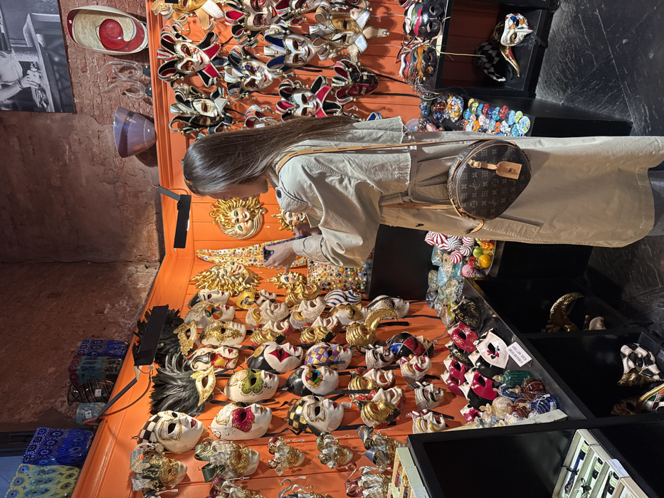 The image depicts an interior view of a retail establishment or gallery in Venice, Italy, characterized by vibrant orange walls densely covered with numerous Venetian carnival masks. A solitary female individual stands facing a corner display, positioned with her back partially to the viewer, holding a dark-colored smartphone vertically to capture a photograph or video.
The individual possesses long, straight, dark brown hair and is dressed in a light-colored, possibly beige or tan, trench coat over dark blue jeans. A round, brown Louis Vuitton crossbody bag with a distinctive monogram pattern and a light-colored luggage tag hangs at her right hip.
The walls are constructed of orange wooden paneling extending from floor level to just below the ceiling, where sections of exposed reddish-brown brick are visible. Mounted black spotlights illuminate the displays. The masks, varying in size and design, include traditional Commedia dell'arte characters, ornate feathered masks, half-masks, full-face masks, and decorative sun-faced masks. Many are intricately painted in gold, white, red, green, and black.
Below the wall-mounted masks, black shelving units display additional merchandise. On the left, shelves hold packaged items and smaller, colorful glass perfume bottles or decorative vials. In the corner, a display features numerous small, brightly colored, spherical objects, possibly glass or ceramic ornaments, along with faux fruit. On the right, shelves showcase more masks, including jester-style and plague doctor designs. The flooring is dark, possibly black or dark grey, with a subtle pattern.