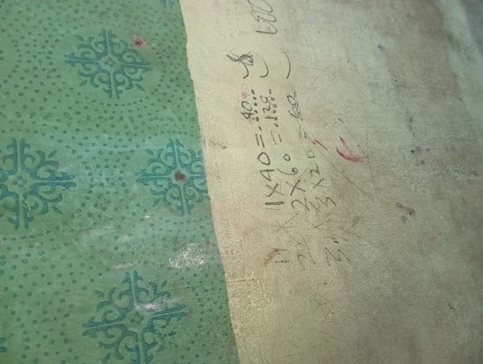 The image shows a close-up view of a wall in Rejosari, Indonesia. The wall is covered in faded green wallpaper with a decorative pattern of swirling motifs and small dots. The lower portion of the wall appears to be a different material, possibly plaster or paint, in a lighter, yellowish-green hue. Scrawled onto this lower section are mathematical equations and numbers written in what appears to be chalk or crayon.
One equation clearly reads "1 x 40 = 40". Below it, "2 x 60 = 120". Further to the right, there is a series of numbers and symbols, some of which are legible like "600". The handwriting is imprecise and seems to be done by a child. There are also some smudges and a faint red mark on the wall, possibly from a crayon or paint. The overall impression is of a worn, interior wall in a residential or educational setting where children may have been present.