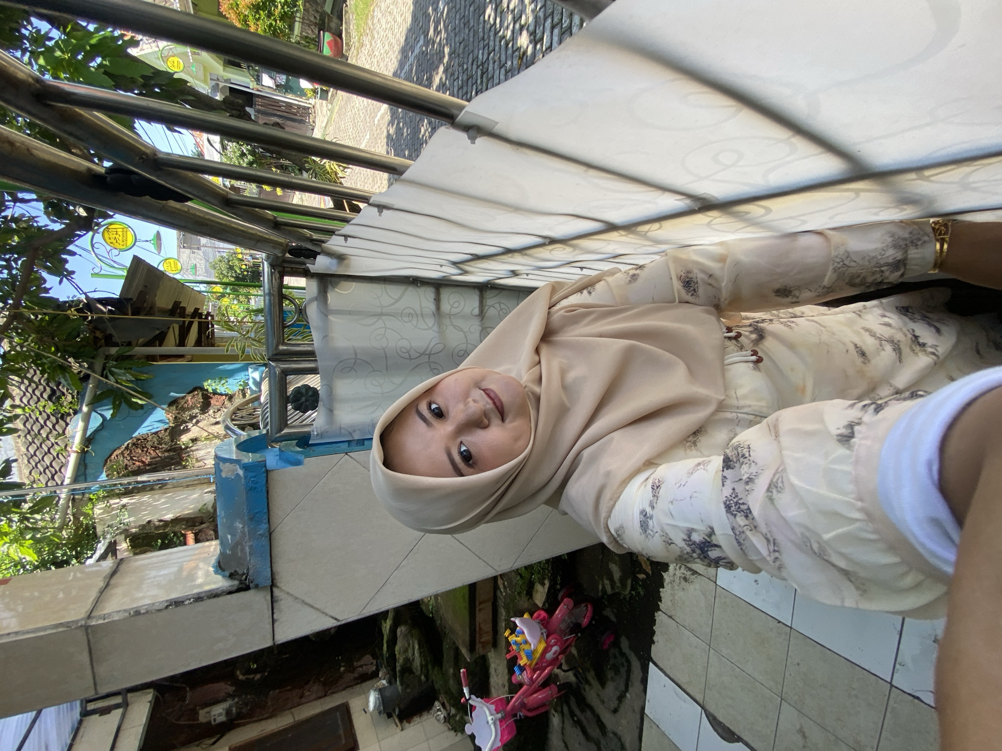 A woman wearing a hijab and a patterned blouse is standing outside in Gelam, Indonesia. She is holding a phone and appears to be taking a selfie. Behind her is a balcony with a metal railing and a translucent roof. In the background, there is a view of a street with houses and trees. Below the balcony, there is a tiled area with a child's pink bicycle.