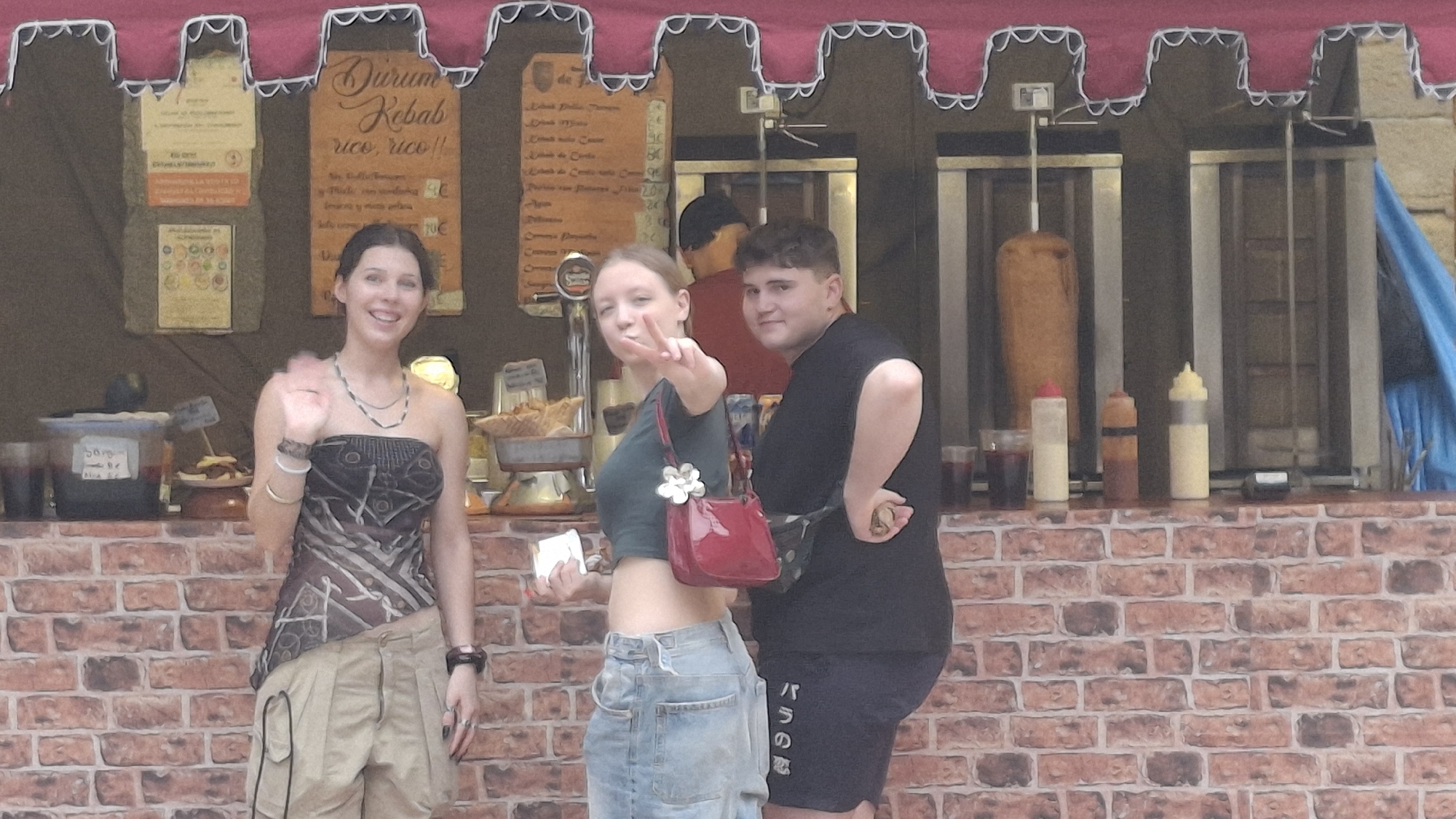 In La Fresneda, Spain, three young adults stand in front of a food stall. A woman on the left, wearing a patterned strapless top and tan cargo shorts, waves with her right hand. To her right, a young woman in a green crop top and denim skirt holds up two fingers in a peace sign and carries a red purse. A young man in a black t-shirt and dark shorts stands to the right of the woman, with his back partially turned. Behind them, a food stall features a vertical rotisserie with meat, condiment bottles, and a brick facade. The stall is decorated with a red awning and hanging signs advertising "Durum Kebab".
