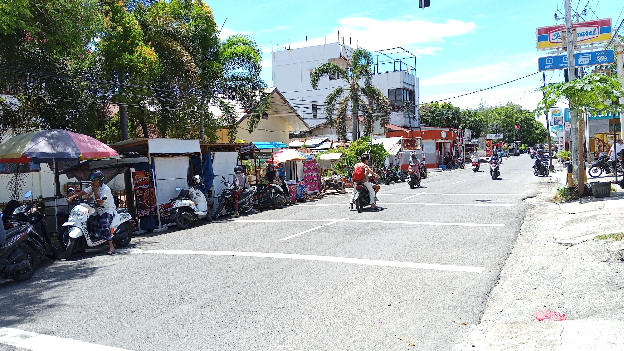 The image depicts a street scene in Pulokiton, Indonesia, on a sunny day with a bright blue sky. A road with white lane markings dominates the foreground, with a concrete sidewalk on the right. Several motorbikes are parked and in motion. On the left side of the road, various small stalls are set up, some with umbrellas providing shade. A man in a white shirt and checkered pants is sitting on a white motorbike in front of one of these stalls. Other motorbikes are parked alongside the stalls, some with riders. In the background, buildings of varying sizes and architectural styles are visible, including a multi-story white building with a rooftop structure. Palm trees and other lush green foliage are interspersed throughout the scene. Several people are riding motorbikes on the road, moving in both directions. A sign for "Indomaret," a convenience store chain, is visible on a pole on the right side of the image, along with other signs advertising services. The overall atmosphere suggests a busy commercial area in a tropical environment.
