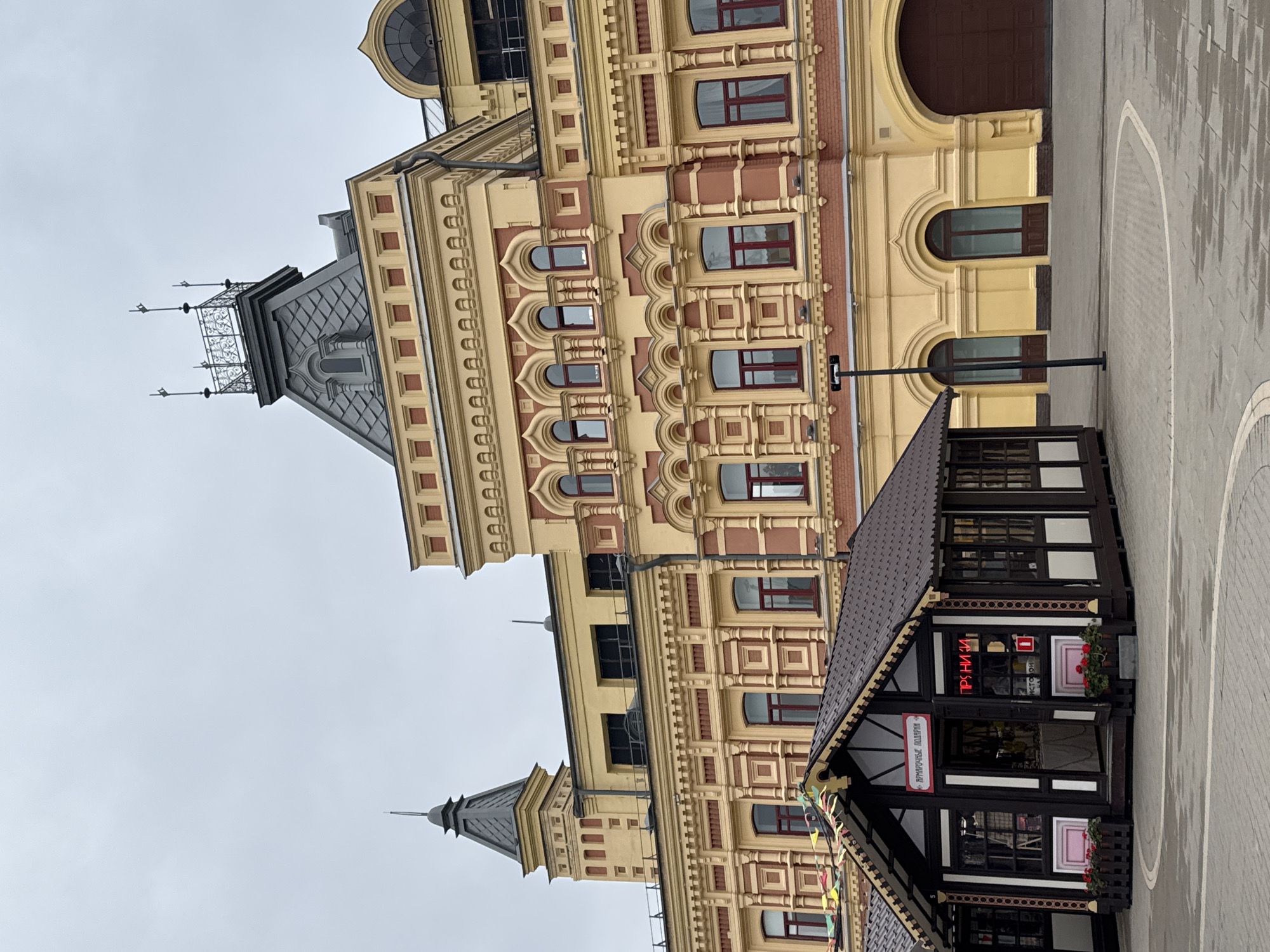 The image depicts a large, ornate yellow and red building in Nizhny Novgorod, Russia, likely an architectural landmark given its detailed facade. In the foreground, a small, dark wooden kiosk with a brown roof is situated on a paved area. The kiosk has large windows and an open entrance, with a sign above it. Red neon lights spell out "ПРЯНИКИ" (Pryanyki, meaning gingerbread or cookies) inside the kiosk. Another sign above the entrance reads "АРМАТОЧНЫЕ ПОДАРКИ" (Armatochnye podarki, meaning "fragrant gifts" or "scented gifts"). Several small flower pots with red flowers are placed in front of the kiosk. The building behind has numerous arched windows with red frames, decorative brickwork, and intricate architectural details. The sky is overcast. The scene appears to be an outdoor market or commercial area within the city.