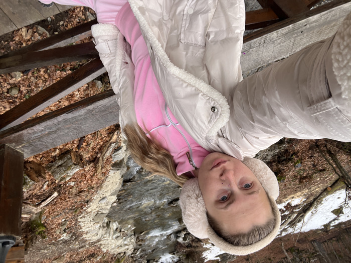A female subject is photographed in a close-up, appearing to take a selfie. She wears a cream-colored puffer jacket with a sherpa-lined collar and cuffs, a pink full-zip tracksuit top, and matching pink tracksuit bottoms. Cream-colored furry earmuffs cover her ears, and her light-colored hair is partially visible over her right shoulder. Her left arm is extended towards the camera's perspective. She stands on a weathered wooden structure, with a railing visible in the foreground, below her. A flowing river with rocky banks is visible behind her, primarily to her right and in the background. The banks are covered with fallen brown leaves. Patches of snow are visible on the left bank and higher ground among deciduous trees, which appear bare. The scene is situated in Crkvine, Montenegro.