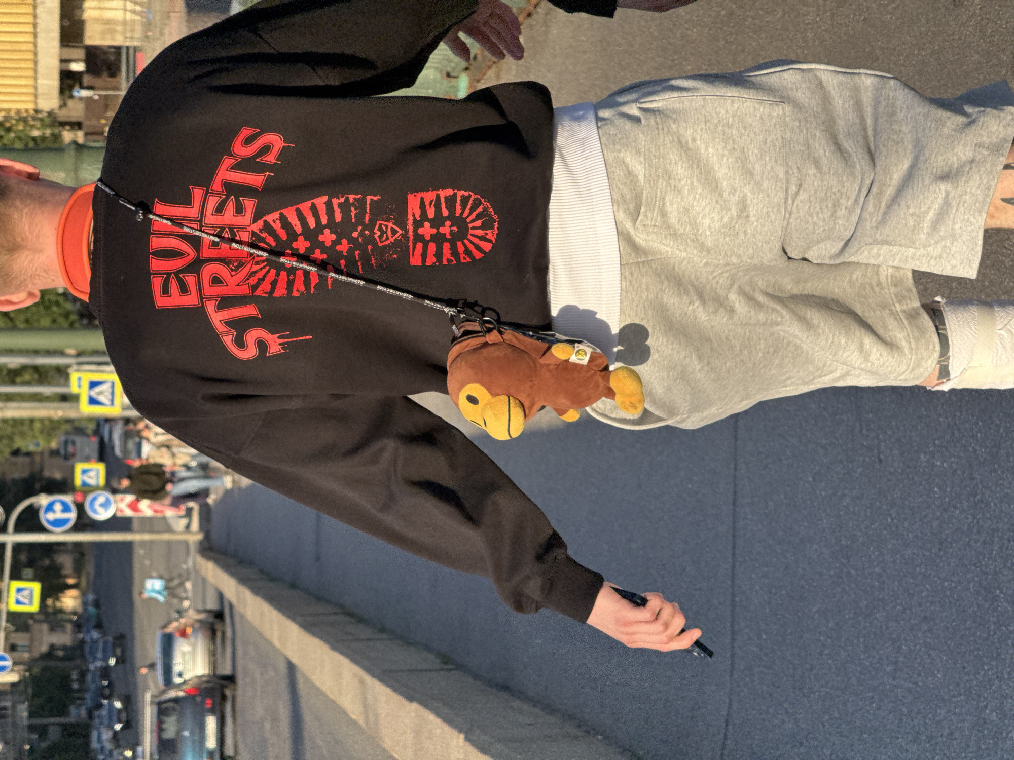 A person is walking on a sidewalk next to a street in an urban environment. The person is wearing a black sweatshirt with red graphic text that reads "EVIL STREETS" and a graphic of a boot sole. A small brown stuffed monkey hangs from a black strap across their chest. The person is also wearing light gray shorts and white socks. In the background, there are cars on the street, buildings, and traffic signs. The lighting suggests it is daytime, likely late afternoon given the shadows.