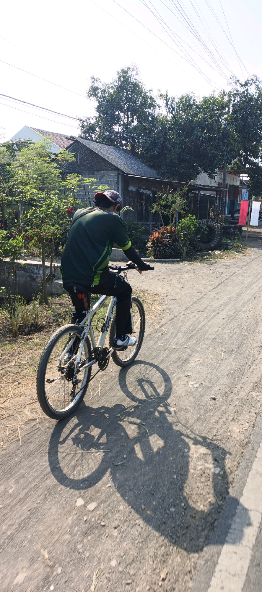 An individual wearing a dark green shirt with green accents and black pants rides a white bicycle on a dirt road in Mojomulyo, Indonesia. The rider is wearing a dark baseball cap and arm sleeves. The bicycle has a dark saddlebag attached to the rear seat post, with a red taillight visible. The background features a house with a tiled roof and grey brick walls, surrounded by lush green trees and vegetation. There are also two flags, one red and one white, planted near the house. The sun casts distinct shadows of the bicycle and rider onto the dusty road. The scene suggests a bright, sunny day in a semi-rural or suburban area.