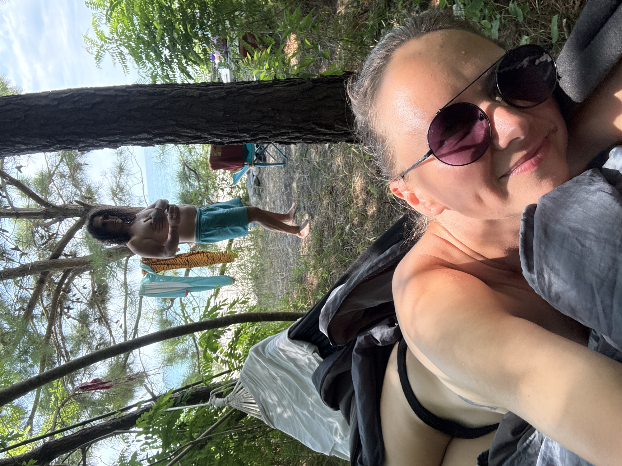 In Yuada-Gulrypsh, Abkhazia, a selfie taken from a hammock reveals a person with sunglasses and dark hair, lying down and smiling. Their head and upper body are in the foreground, with a black bikini bottom visible. In the background, a second person stands shirtless, wearing blue shorts, with several items of clothing hanging on a line between trees. The scene is set outdoors, near a body of water, with trees and foliage surrounding the area. The lighting suggests it is daytime.