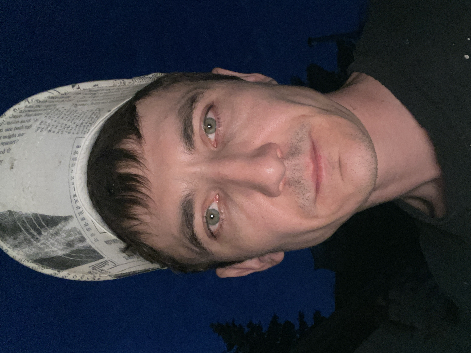 A close-up, frontal, head-and-shoulders shot shows a man outdoors at dusk, wearing a novelty hat made of newspaper. The man has short, dark hair, light green eyes, and a light complexion. He is wearing a dark t-shirt. The background is dark, with a hint of trees on the left and a dark sky. The image is taken in Laishevo, Russia.