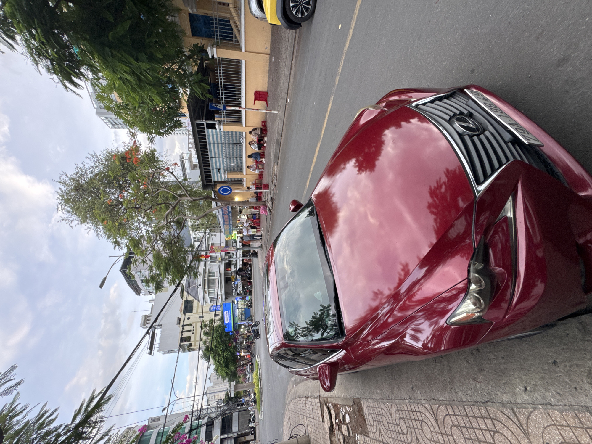 The image shows a street scene in Nha Trang, Vietnam. A dark red Lexus sedan is parked on the side of the road, facing forward. The car is in focus in the foreground, with its hood and windshield reflecting the sky and surrounding trees. The street is paved with asphalt and has a yellow dividing line. To the left of the car is a sidewalk with a patterned concrete surface, and beyond that, a narrow patch of dirt with some debris.
In the background, there are several buildings of varying heights, including a multi-story modern building with a glass facade and an older building with a yellow exterior. Trees with green leaves and some with red flowers are visible along the street and in front of the buildings. A power line with many cables stretches across the upper part of the image.
On the sidewalk, several people are seated at small tables, some of them eating. There are red plastic stools and a few umbrellas providing shade. A blue traffic sign with a white circle and arrow is present. Further down the street, there is a rotary intersection with traffic, including motorcycles. A yellow SUV is partially visible on the right side of the image, parked behind the Lexus. The sky is partly cloudy, suggesting it is daytime.