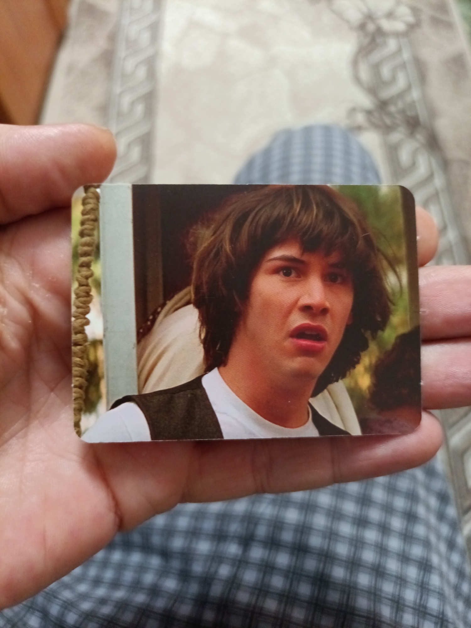 A close-up, eye-level shot captures a hand holding a rectangular, glossy photograph. The photograph features a young man with shaggy brown hair, a white t-shirt, and a dark vest, his mouth open in a look of surprise or shock. The background of the photograph is blurred, suggesting an outdoor or dimly lit setting. The hand holding the photograph is an adult's, with visible skin texture and fingernails. The image is taken against a backdrop of a patterned fabric, possibly upholstery or clothing, with a plaid design in shades of grey and white. There is no discernible information about the location in the city of Nyagan, Russia.