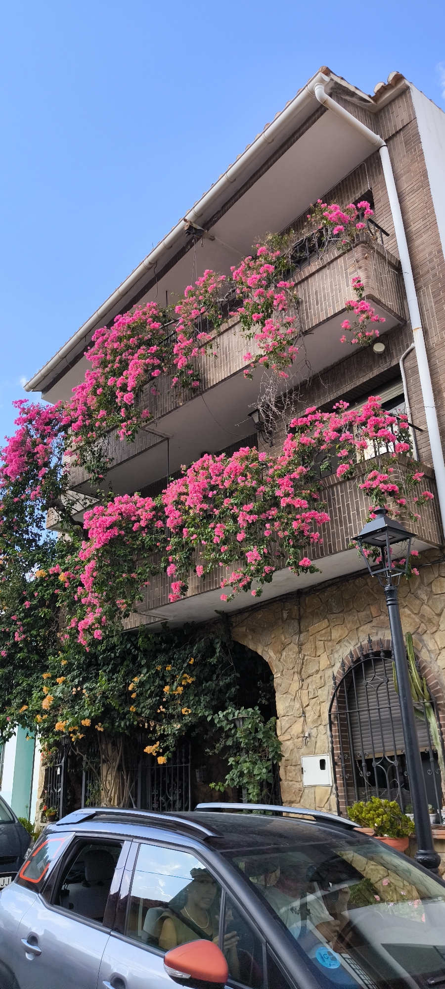 This image shows a three-story building with balconies overflowing with vibrant pink bougainvillea flowers in Fanzara, Spain. The building features a brick facade and a stone-arched entrance on the ground floor. A black lamppost stands to the right of the entrance. In the foreground, a silver car with a black roof is parked, partially obscuring the lower part of the building. Two people are visible inside the car. The sky is clear and blue.