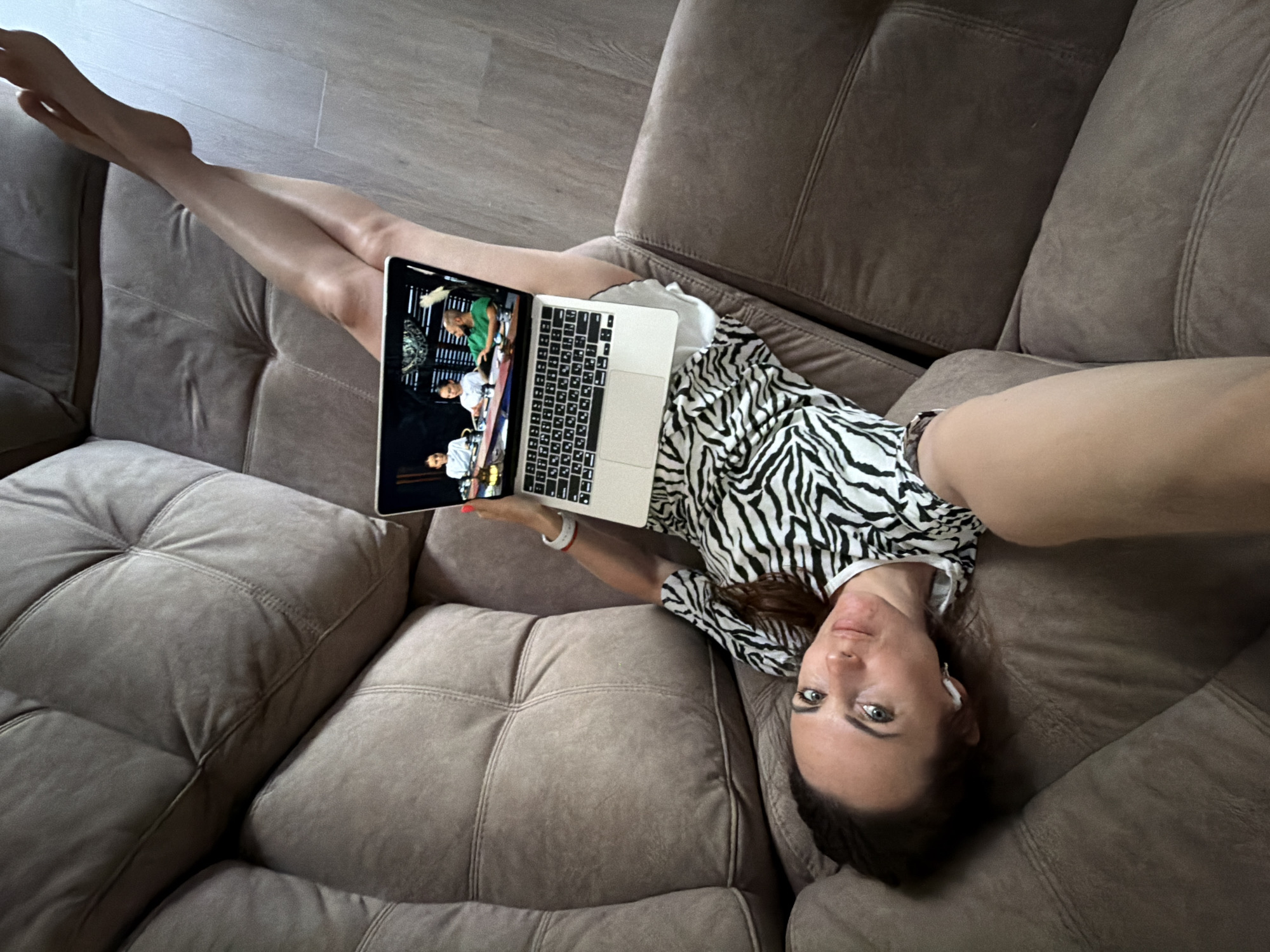 A woman lies on a brown sectional sofa, holding a laptop. The laptop screen shows a video of people, one wearing a green shirt. The woman is wearing a black and white zebra-striped top. She has dark hair, light skin, and is wearing an earpiece. She is looking towards the camera with one arm extended, likely taking a selfie. The location is in Birobidzhan, Russia. The sofa is large and sectional, with a tufted design on the cushions. The flooring is wood-toned.