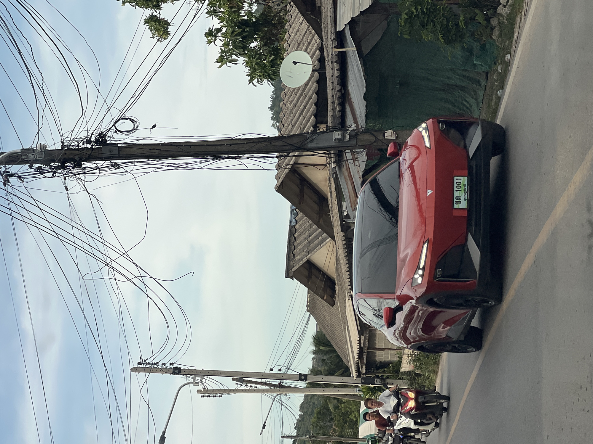 The image depicts a street scene in an unspecified city, Thailand. The street has a red car parked with the license plate "ขต 1001". The car has a black roof, red side mirrors and is parked on the right side of the street. The street also has two motorbikes and riders are traveling on it. Two riders are wearing white shirts. Above the street, there are utility poles burdened with a tangle of wires. Buildings with corrugated metal roofs and a satellite dish are situated alongside the street. The sky is overcast.