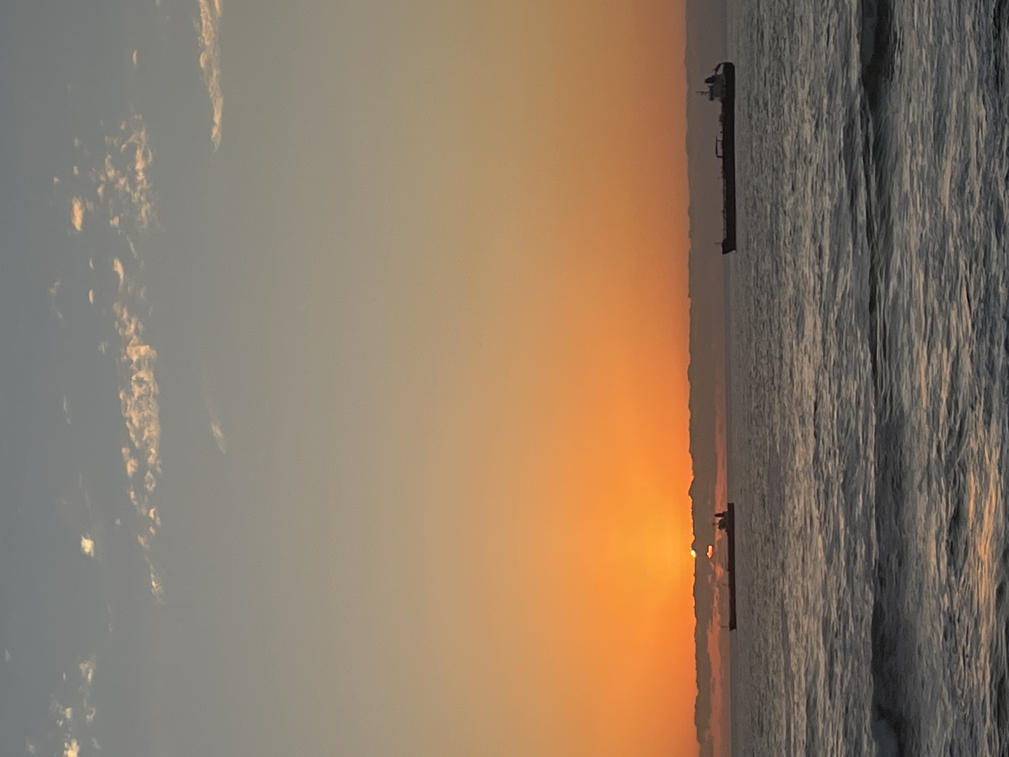 The image depicts a sunset over the Pacific Ocean in Arica, Chile. The sun is setting near the horizon, casting an orange and yellow glow across the sky and reflecting on the water. A cargo ship is silhouetted against the setting sun in the distance. There are few clouds in the sky. The ocean's surface appears choppy, with small waves visible.