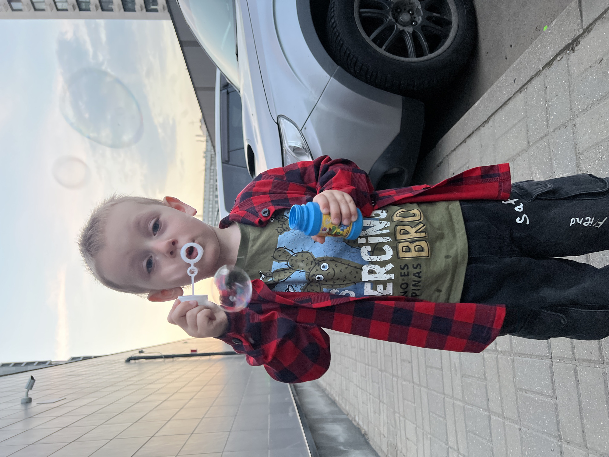 A young boy stands on a sidewalk in what appears to be an urban setting. He is blowing bubbles using a wand and a bottle of bubble solution. He wears black jeans with white text, a green t-shirt with a print and a red and black plaid shirt. A silver car is parked behind him, and a large building is in the background. The scene takes place outside at dusk or dawn. The setting is Mytishchi, Russia.