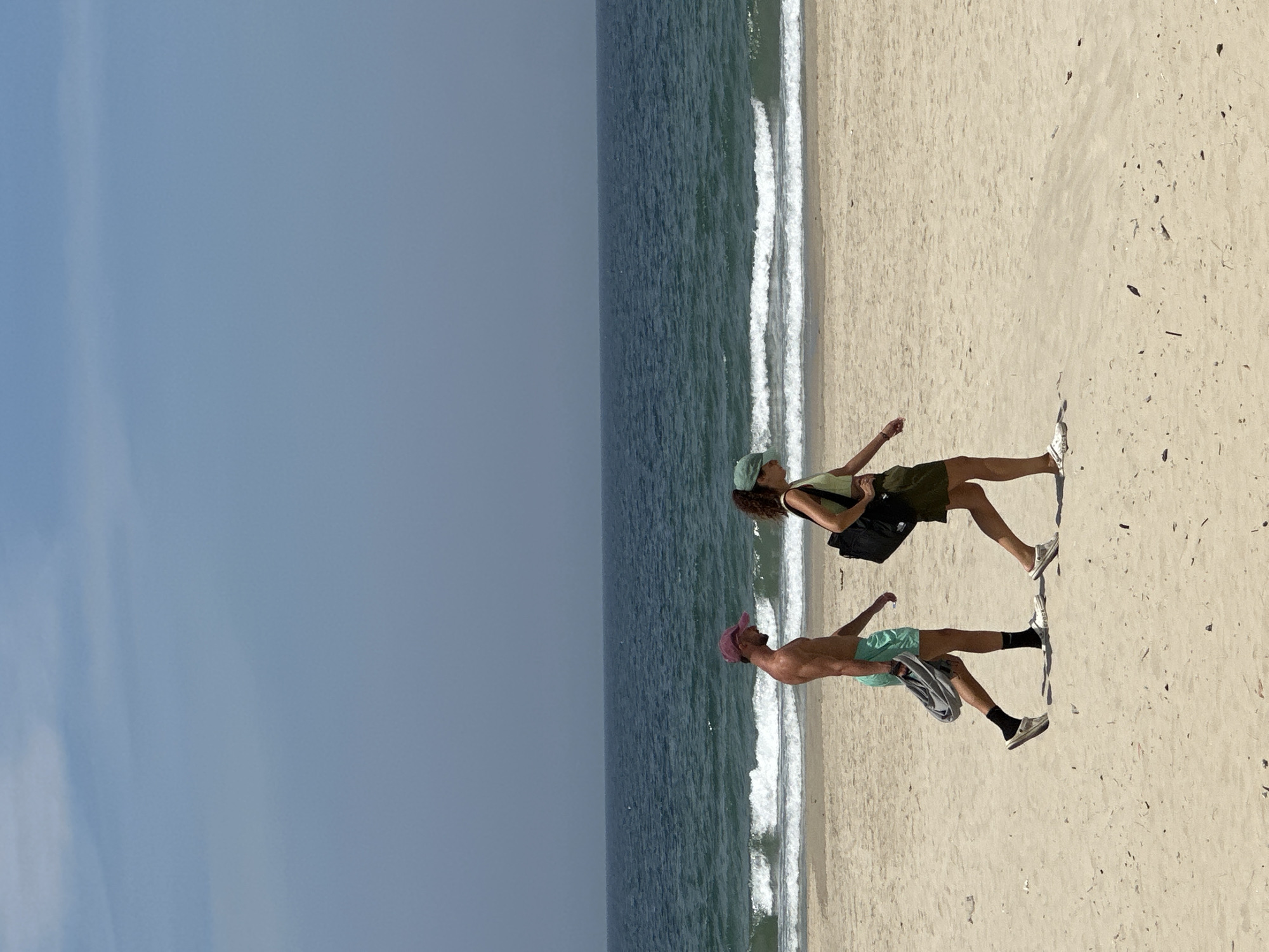 Two individuals, a man and a woman, are walking on a sandy beach. The man, on the left, is wearing teal swim shorts and a pink baseball cap. He has a muscular build and is carrying a folded gray towel in his left hand. The woman, on the right, is wearing a light green tank top, a dark green skirt, and a green baseball cap. She is carrying a black bag over her right shoulder. Both are wearing casual footwear, appearing to be slip-on shoes. They are walking from left to right, parallel to the ocean. The ocean is visible behind them, with gentle waves breaking on the shore. The sky above is a clear, light blue, with some faint clouds. The scene is set on a sunny day at a beach, likely in the city of Da Nang, Vietnam, known for its beautiful coastline.