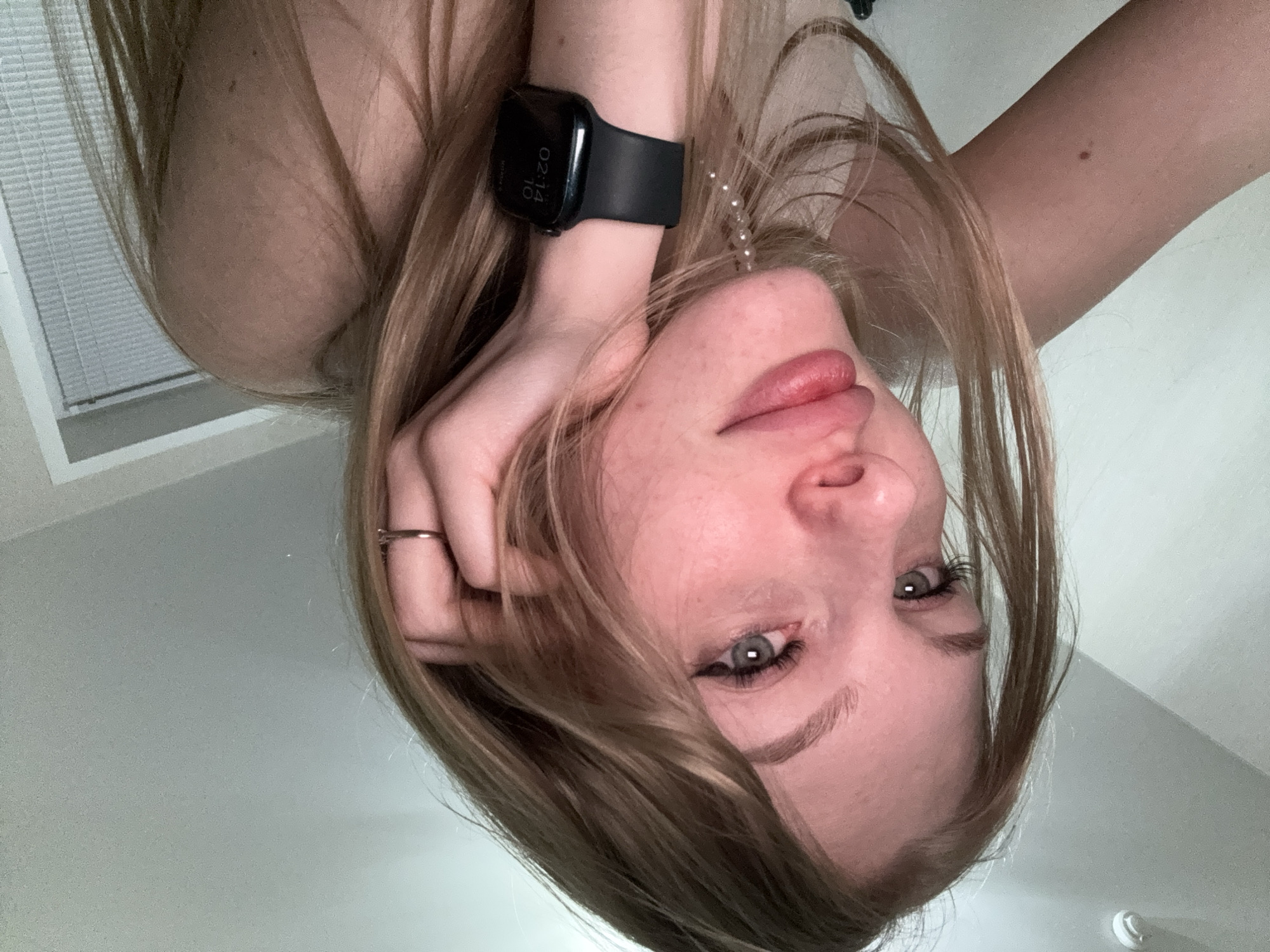 A close-up selfie of a young woman with blonde hair, wearing a black smartwatch on her left wrist. She has blue eyes, winged eyeliner, and full lips. Her right hand is resting on her face, and she is wearing a ring on her right ring finger. A pearl necklace is visible hanging from her chest. The background appears to be an indoor setting, possibly a room, with a plain white wall and a window with blinds. There is no indication of the city of Perm, Russia, in the image.